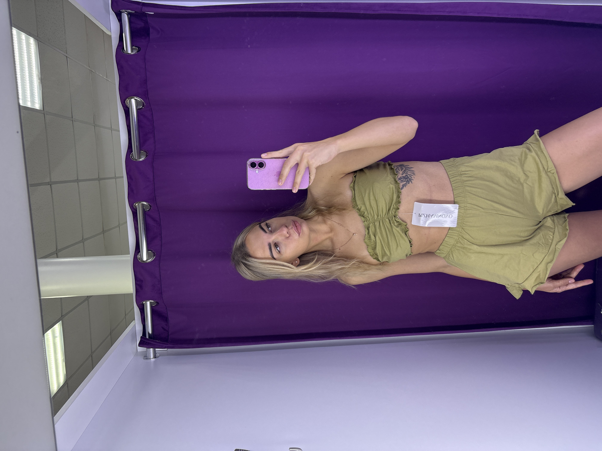 A young woman with blonde hair is taking a selfie in a fitting room. She is wearing a green strapless top and matching shorts. The fitting room has a purple curtain and white walls. A tag is hanging from her top. The background shows a ceiling with fluorescent lighting.