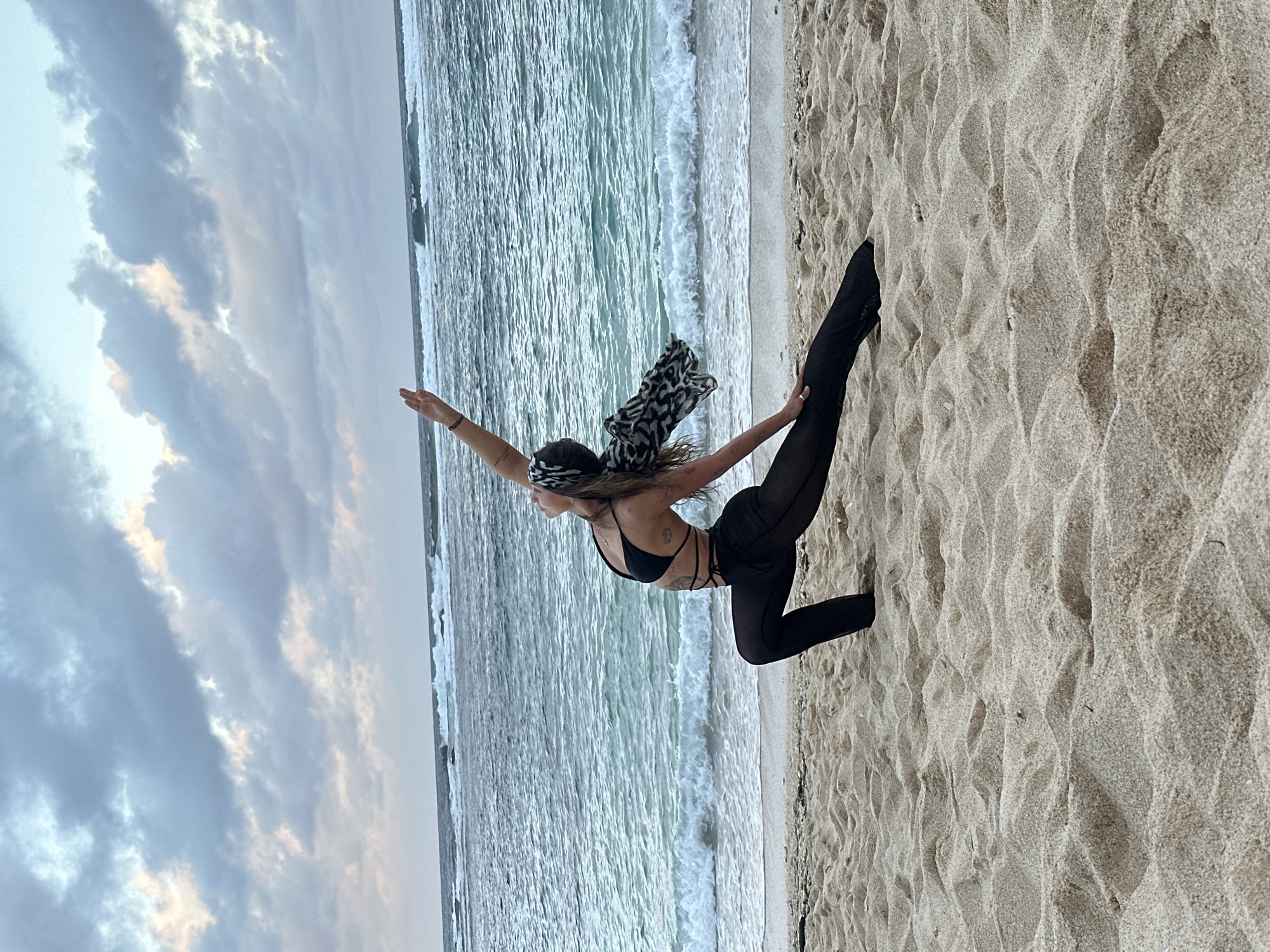 A woman in a black bikini, sheer black pants, and a patterned headscarf stands on a beach in Kuta Selatan, Indonesia. She faces the ocean with her arms raised, striking a yoga-like pose. The sky is cloudy, and gentle waves wash onto the sandy shore.