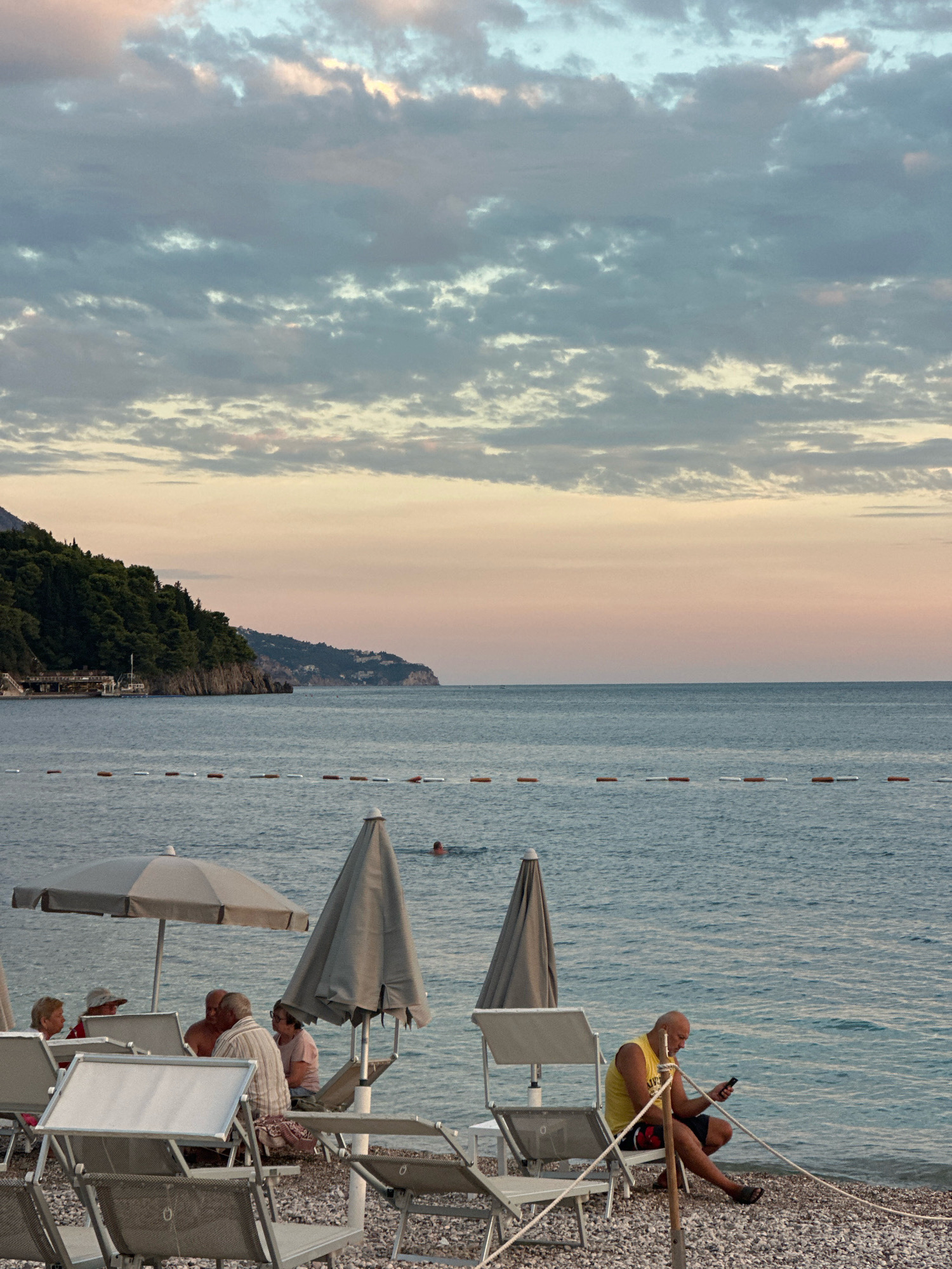 The image depicts a serene beach scene in Budva, Montenegro, likely during the late afternoon or early evening. The sky is filled with soft, pastel-colored clouds transitioning from pale pink to a muted blue, indicating either sunrise or sunset. In the foreground, several empty lounge chairs and umbrellas are arranged on a pebble beach, suggesting a quiet moment before or after peak visitor hours.
Several individuals are present. To the left, a group of people are seated together under a large umbrella, appearing to be engaged in conversation. Further to the right, a lone man is sitting on a chair near the water's edge, focused on his mobile phone. His posture is relaxed, and he is dressed in a yellow sleeveless shirt and dark shorts. In the background, the calm sea stretches to the horizon, with a buoy line marking a boundary in the water. A lone swimmer can be seen in the distance. The coastline is lined with lush green trees and rocky formations, characteristic of the Budva Riviera's landscape. Buildings and structures are visible on the hillsides in the distance, hinting at the town of Budva. The overall atmosphere is one of peaceful relaxation and coastal beauty.