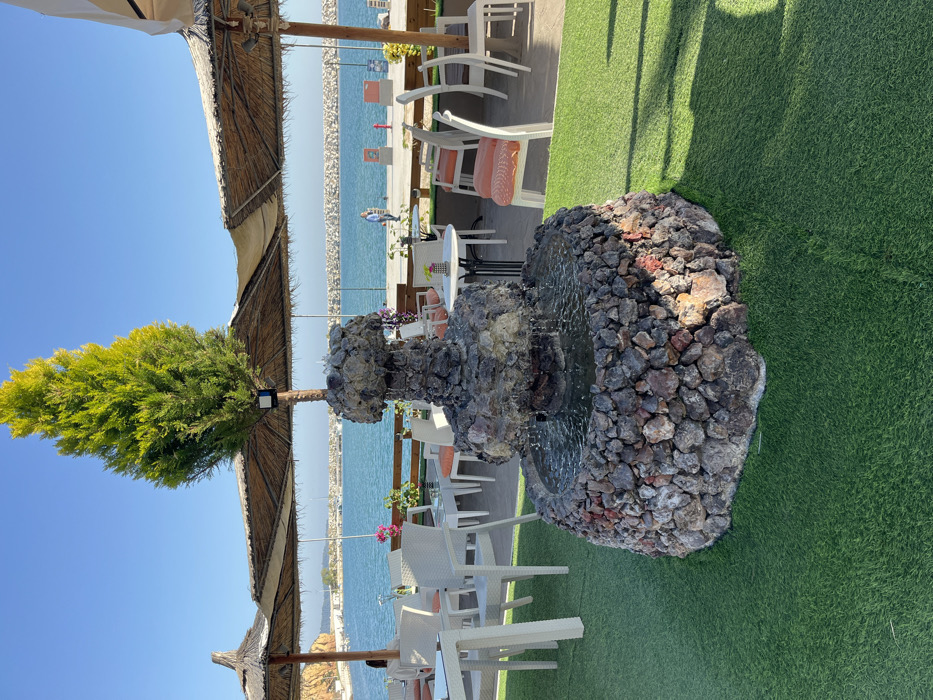 The image shows a stone fountain with water cascading down its tiers in the center of a patio. The patio is surrounded by a green, artificial grass surface and is partially shaded by a brown thatched roof. White tables and chairs are set up around the fountain. The background features a clear blue sky, a blue body of water, and a row of white rocks lining the water's edge. One person is visible in the background, walking towards the right. The image was taken at latitude 40.626 and longitude 24.576, which corresponds to a location in Limenaria, Greece.