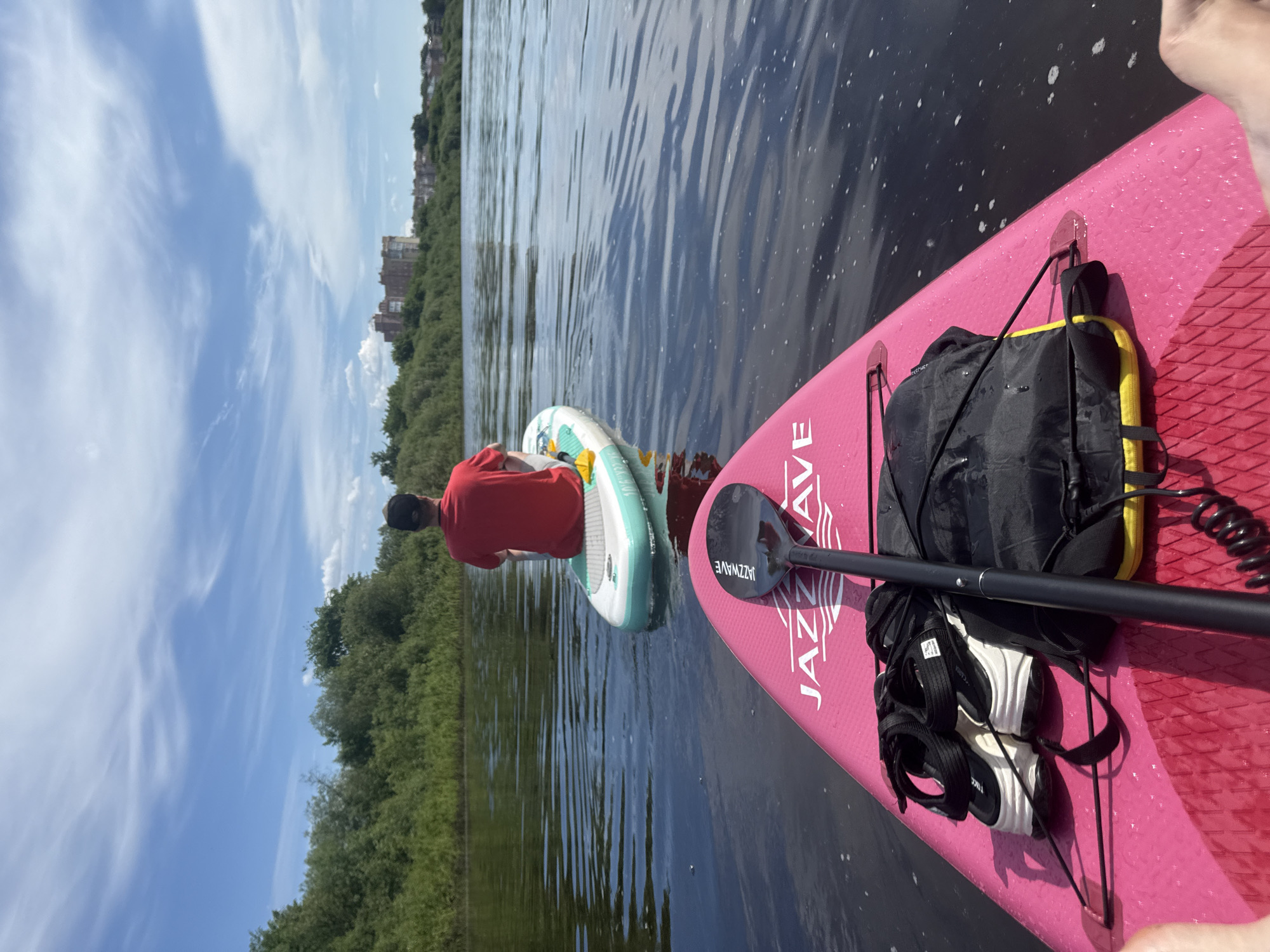 The image captures a first-person perspective from a pink inflatable paddleboard on the Vologda River in Vologda, Russia, under a clear blue sky with sparse, wispy clouds. In the immediate foreground, the textured surface of the pink paddleboard displays a black paddle, a pair of black and white chunky-soled sandals, and a black waterproof bag with a yellow bottom, all secured. Ahead and slightly to the right, a second individual, viewed from the back, sits on a light blue and white inflatable paddleboard. This person wears a red t-shirt and a dark baseball cap. The river's surface is dark, calm, and shows minor ripples. Both riverbanks are densely vegetated with various green trees and bushes. On the right bank, behind the treeline, several multi-story buildings, consistent with an urban setting, are visible, including one prominent, older-style structure.