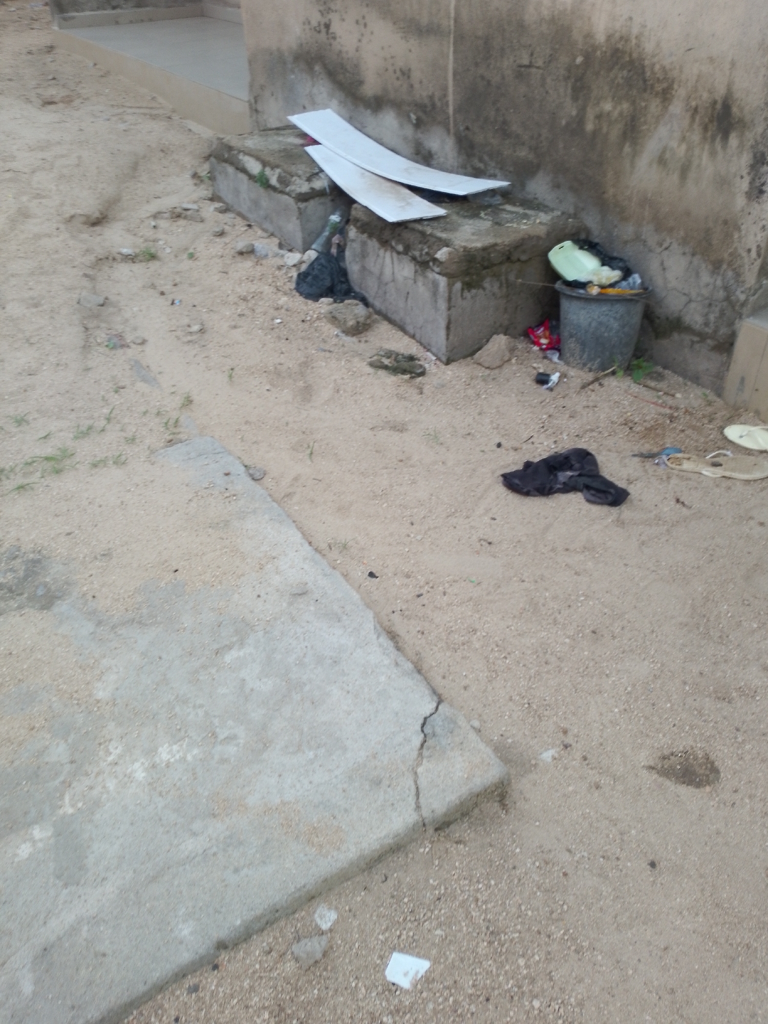 The image depicts an outdoor scene with a sandy ground and a concrete structure. In the background, a weathered wall runs horizontally. On the ground, a gray trash can is filled with various items, including a light green lid and black trash bags. Nearby, a pile of black clothing is scattered on the sand. To the left, a section of a concrete structure with a cracked surface is visible. Two pieces of white material, possibly cardboard or plastic, are placed on top of a concrete ledge in the mid-ground. The scene appears to be in an outdoor urban or semi-urban environment.
