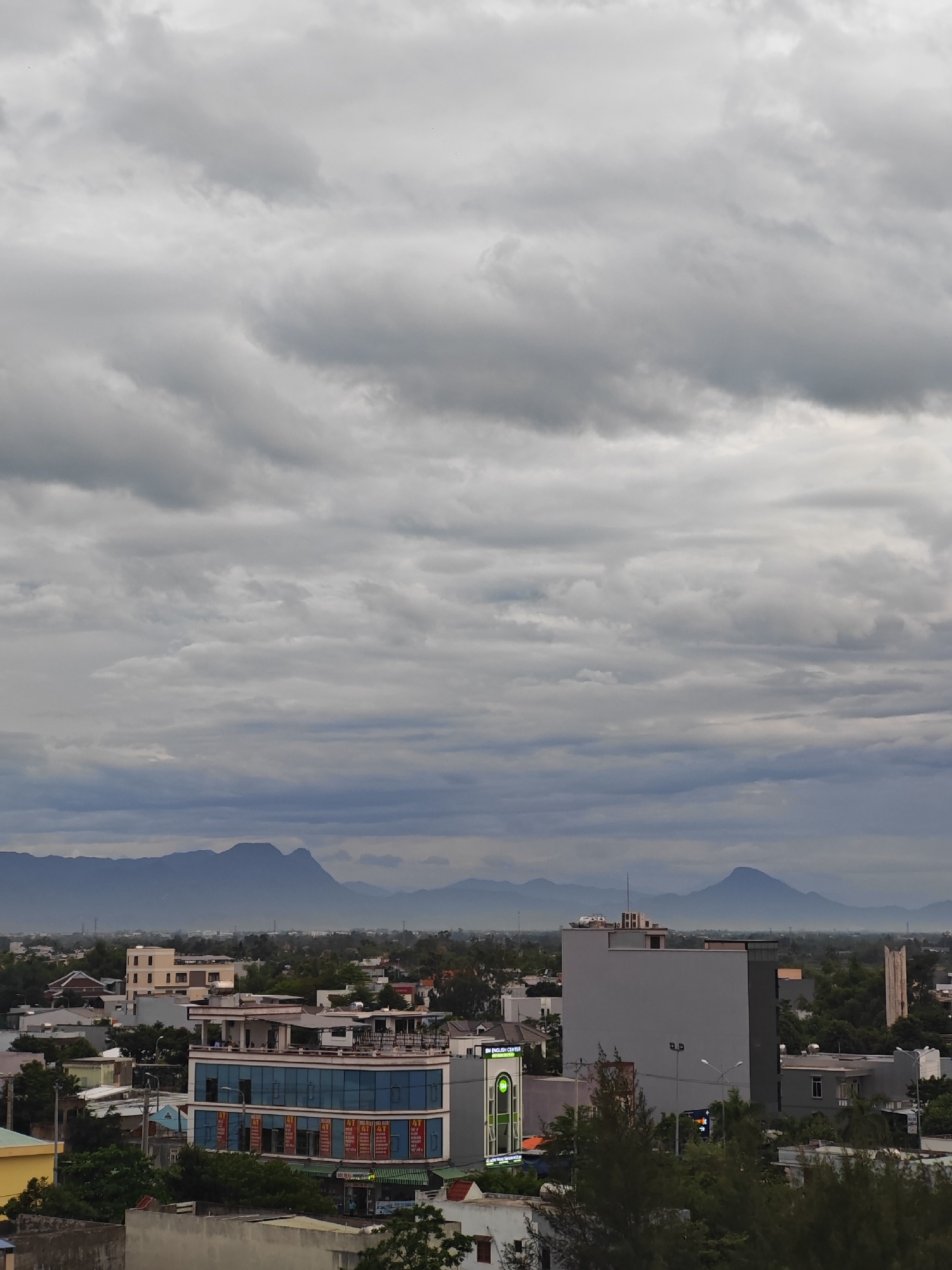 This is an outdoor, eye-level, vertical, full shot of a city skyline in Da Nang, Vietnam, with a mountain range in the background under a cloudy sky. In the foreground, there are several buildings, some with blue glass facades and advertisements on them. Trees and other smaller structures are visible throughout the scene. The mountains in the distance are hazy and appear to be covered in mist. The overall mood of the image is somber and overcast, suggesting a cloudy or potentially rainy day.