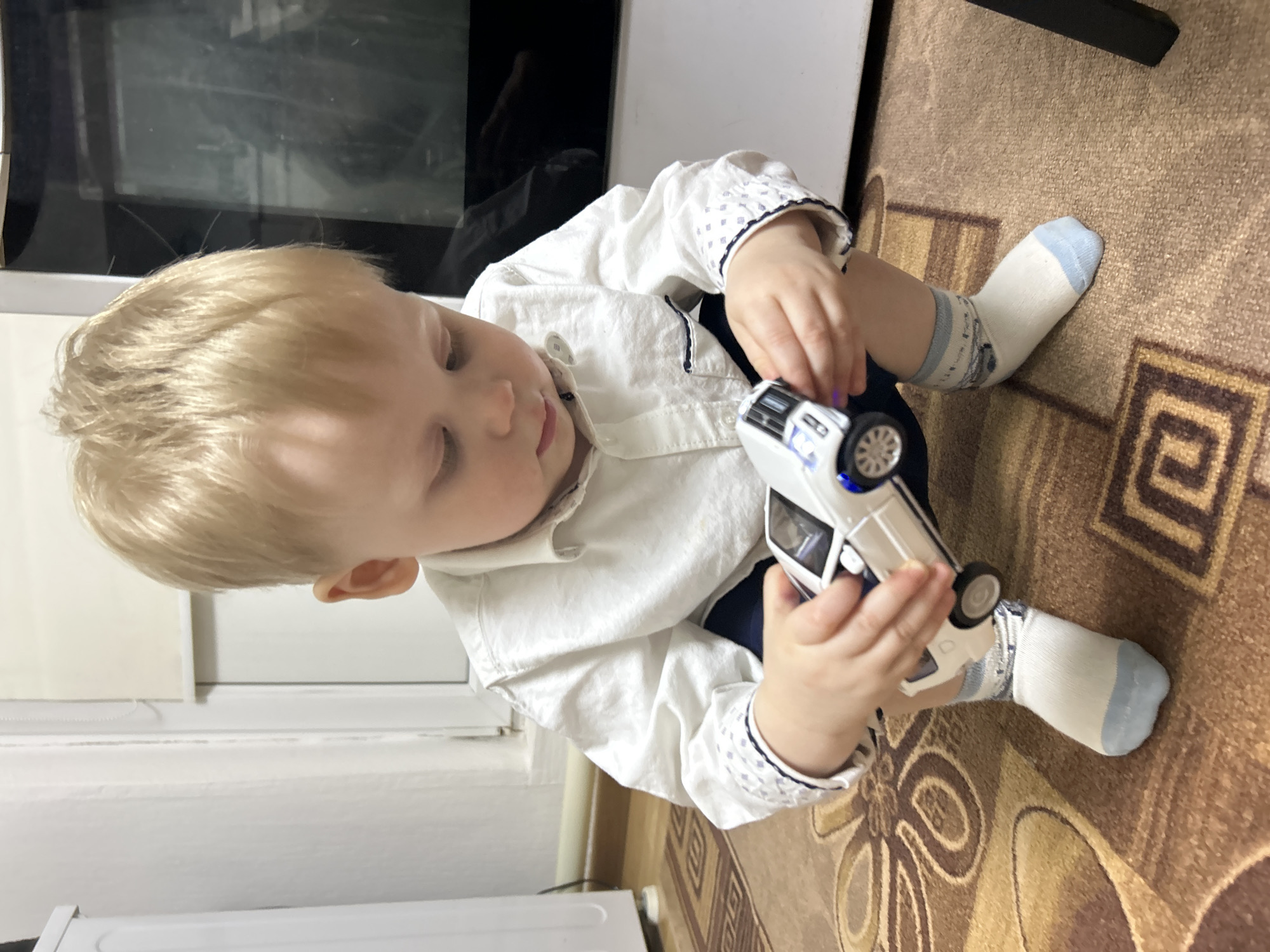 The image displays a young child, approximately a toddler, with short blond hair, in a crouching position on a patterned carpet indoors. The child wears a white long-sleeved collared shirt with dark blue piping and patterned cuffs, dark blue shorts or pants, and white socks with light blue heels/toes. Both hands hold a white toy vehicle, possibly an SUV or police car, with an illuminated blue light visible near the front wheel well. The child's gaze is directed downward at the toy. The immediate environment includes a brown carpet featuring a repeating geometric design in shades of brown and beige. In the background, to the left, a white vertical appliance or piece of furniture is partially visible, with a dark cable running along a white baseboard. To the right, a large dark, reflective surface, likely an appliance door (such as an oven), is visible, showing a faint, inverted reflection. A white window or door frame with a blind is present in the central background. The specific city of Фоки, Russia, cannot be visually determined from the content of this indoor residential scene.