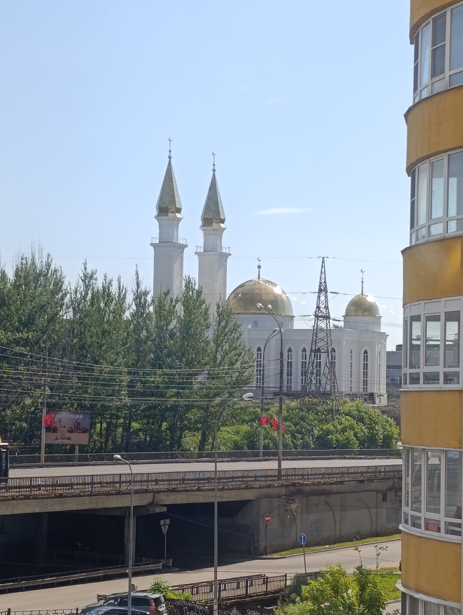 The image captures a street view in Penza, Russia, featuring the Penza Mosque as a prominent landmark. The mosque is characterized by its white facade, two tall minarets topped with crescents, and two golden domes. In the foreground, a multi-level highway overpass with concrete supports dominates the scene. A yellow, modern apartment building with rounded balconies is visible on the right side of the frame. Lush green trees line the area behind the overpass and around the mosque. A large advertisement billboard is affixed to the side of the overpass. The sky is clear and blue, indicating a daytime setting. There are no visible people in the frame.