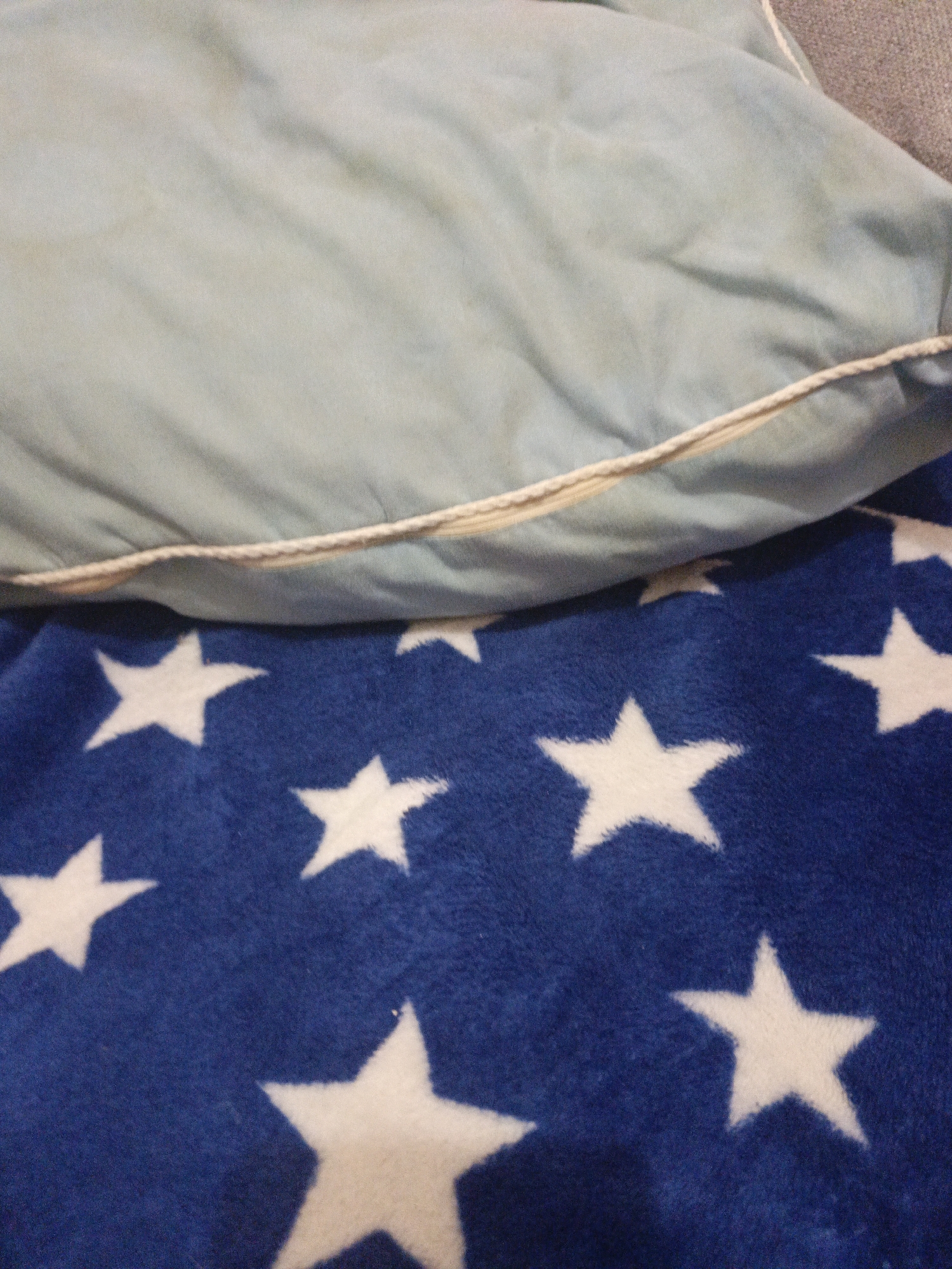 The image shows a close-up view of bedding. The upper portion of the image features a light blue, wrinkled fabric, possibly a pillowcase or blanket, with a cream-colored cording detail visible along its edge. Below this, a dark blue fabric with a pattern of white stars is prominent, extending across the lower half of the frame. The stars vary slightly in size and orientation. There is no visible indication of the city of Uren, Russia, or any people or actions within the scene.