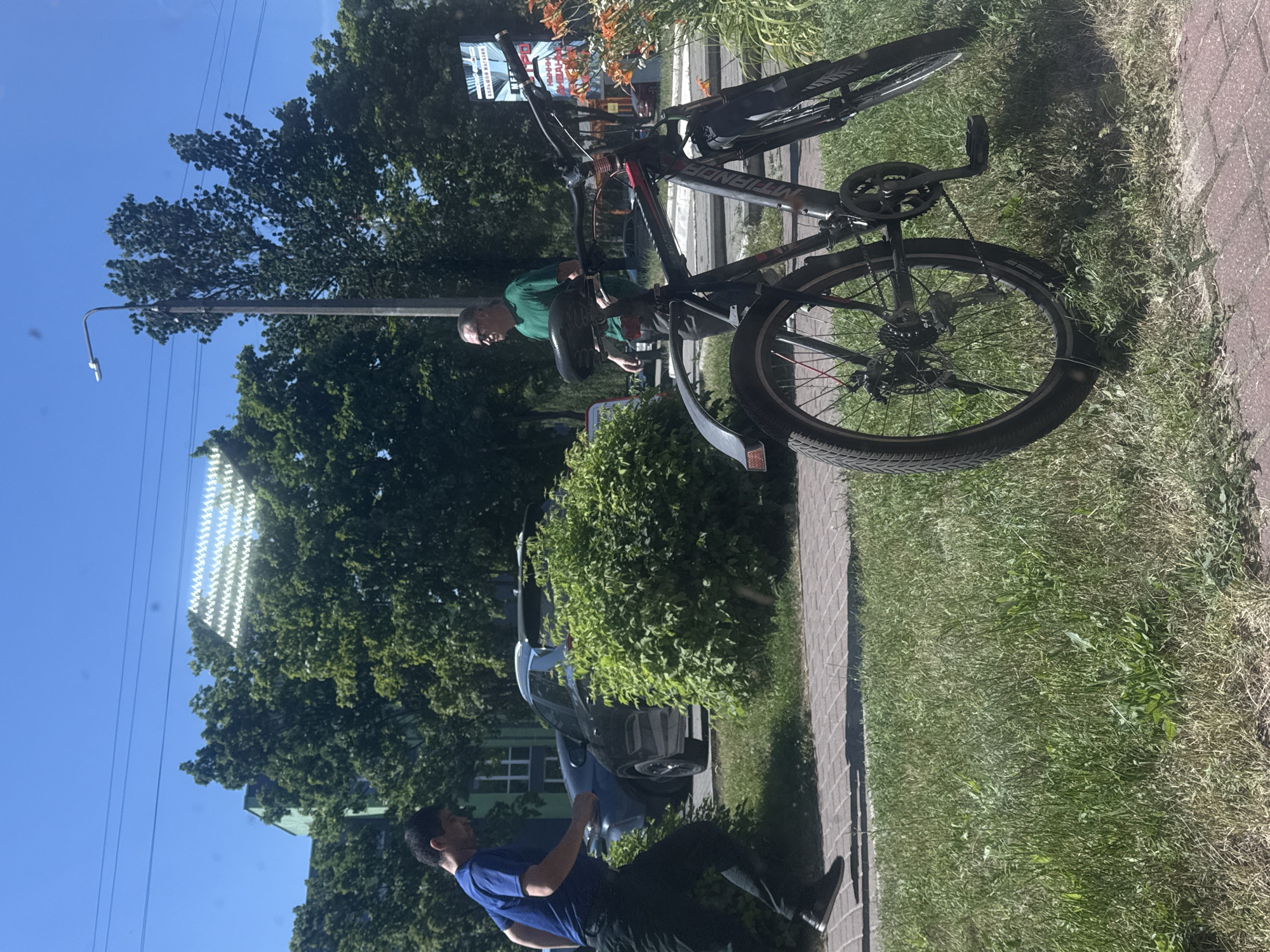 The image depicts an outdoor scene, likely in Barysaw, Belarus, on a sunny day. The primary focus is on a man in a green t-shirt standing next to a black bicycle. The bicycle is a prominent object, with its frame, wheels, and handlebars clearly visible. The man appears to be interacting with the bicycle, possibly in the process of working on it or preparing to ride.
In the background, lush green trees suggest a temperate season. A light pole with a streetlamp is also visible, indicating an urban or suburban setting. Further in the distance, a dark-colored car is parked near a building with green window frames. Another person, wearing a blue t-shirt and black pants, is seen in the lower part of the image, facing away from the camera and appearing to be walking.
A paved pathway runs alongside the grassy area where the bicycle and the man in green are located. There is a sign in the background with some text, but it is not clearly legible due to its distance and resolution. The overall impression is of a casual daytime activity taking place outdoors. The bright sunlight and clear blue sky indicate good weather conditions. The presence of trees, a streetlamp, and a car suggests a location within a town or city.