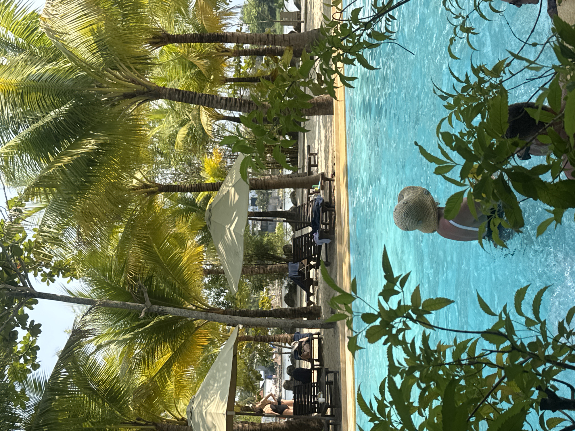 The image shows a swimming pool area with palm trees and lounge chairs. Several people are relaxing on the chairs under white umbrellas, while a woman wearing a hat and swimsuit is in the pool. The location appears to be a resort or hotel pool area.