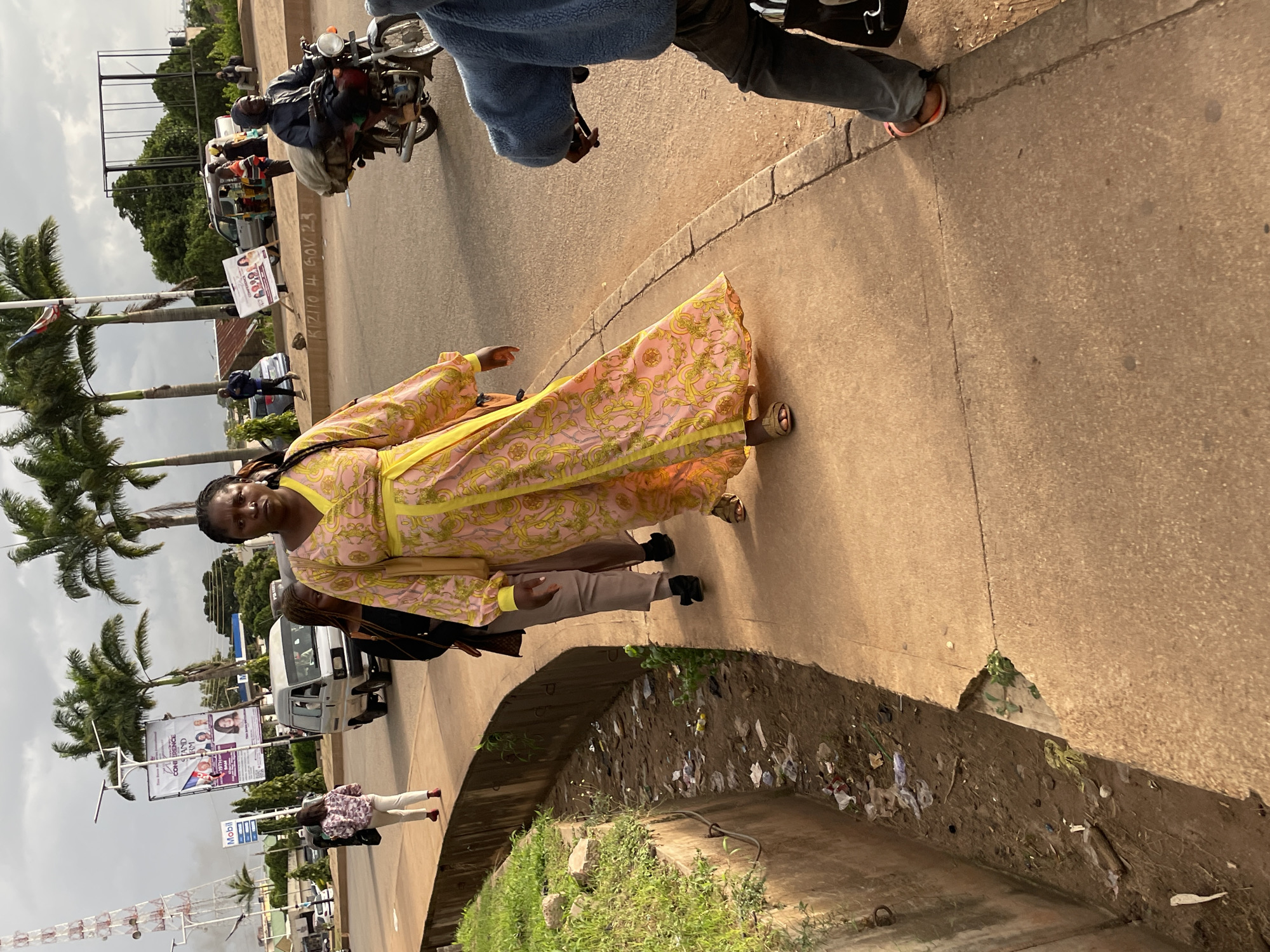 This image depicts a street scene in Jos, Nigeria, on an overcast day. A woman in a vibrant yellow and pink patterned dress walks down a concrete sidewalk. She has dark skin and is looking down. Behind her, another woman in beige pants and a dark top walks, also with dark skin. A third person, mostly obscured, is walking behind them in dark clothing.
The sidewalk runs alongside a drainage ditch filled with dirt, debris, and some scattered garbage. Beyond the sidewalk, a paved road is visible, with vehicles including a white van and a motorcycle with a rider carrying a large sack. Several palm trees are scattered throughout the background, along with a tall communication tower and various buildings. Advertisements are posted on poles near the road. The overall impression is that of an urban environment in Nigeria, likely during daylight hours, with a mix of pedestrian and vehicular activity.