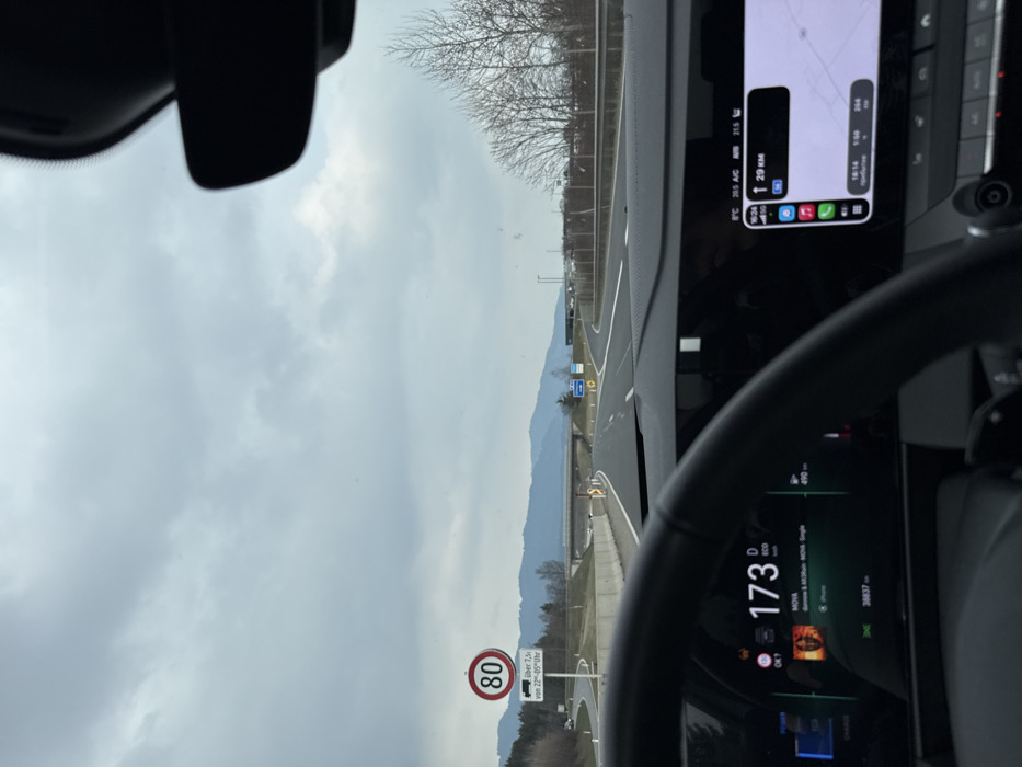 The scene is viewed from inside a vehicle on a multi-lane highway near Aichdorf, Austria, under an overcast sky. The lower left of the frame is occupied by a dark steering wheel. The digital instrument cluster behind it displays a speed of 173 km/h, 'D' for drive mode, 'ECO' indicator, and an odometer reading of 38,837 km. An album cover for "MOVA" by "dannn & M.M.Run" is visible, indicating music playback from an iPhone. To the right, an infotainment screen shows a navigation map with 29 km remaining to the destination, the current time as 16:24, and an outside temperature of 8°C. On the left side of the highway, a circular speed limit sign displaying "80" is present, with a supplementary sign stating "über 7.5t von 22-05 Uhr" (for vehicles over 7.5 tons from 10 PM to 5 AM). The highway, marked with white lines, extends into the distance, passing beneath a concrete overpass. Distant mountains are visible on the horizon, and bare deciduous trees line the right side of the road. No individuals are visible.