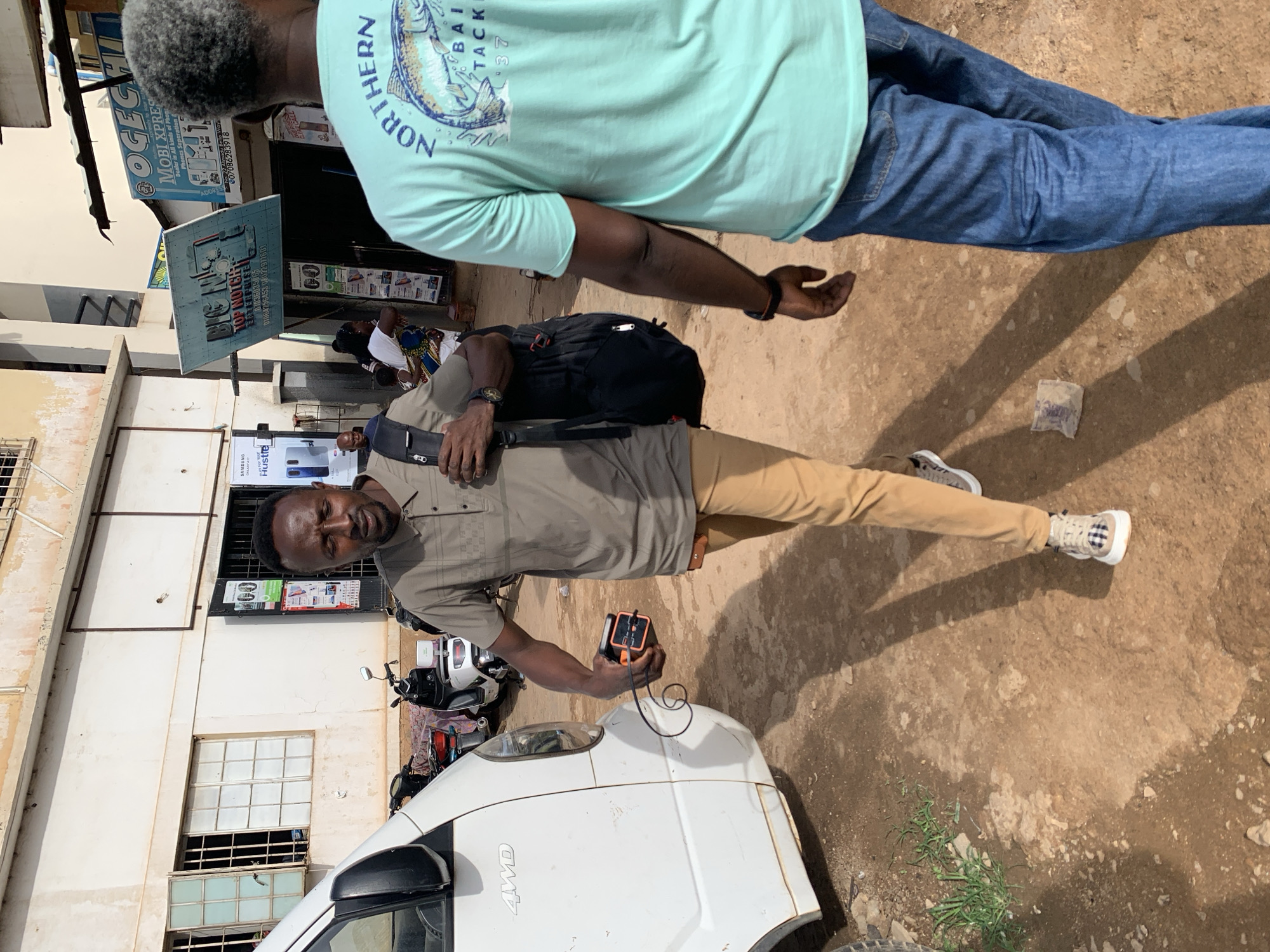 An outdoor scene in what is likely Jos, Nigeria, shows a man holding an electronic device near the front of a white 4WD van. The man is wearing a khaki-colored shirt and pants, with a black bag slung across his shoulder. Another man, partially visible, is wearing a light green shirt and blue jeans. A sign reads "OGEGHI MOBI XPRES", and mentions Samsung Galaxy A1 along with phone numbers. There are also motorcycles parked nearby and people sitting by a building in the background. The ground is dirt or gravel.