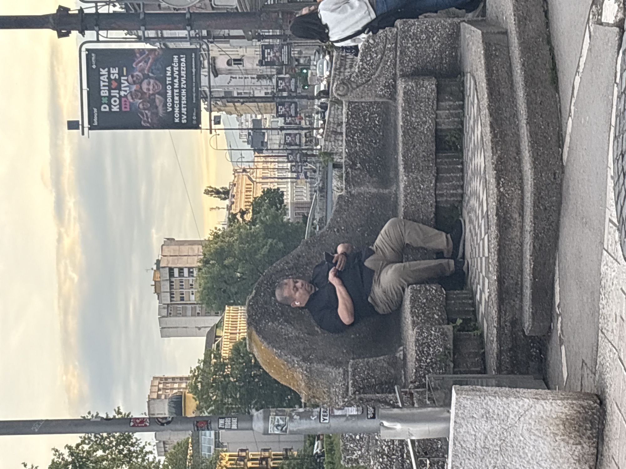 The image shows a man relaxing on a stone structure that borders a set of stairs in Sarajevo, Bosnia and Herzegovina. He is lying on his left side, wearing a black shirt, khaki pants, and black shoes. He has an earbud in his left ear and is holding a phone in his hand. The stairs are made of gray stone. Behind the man are buildings, trees, and a street with cars. There are advertisements on the street poles. One advertisement is for a concert promotion. The sky is overcast, suggesting it is either early morning or late afternoon.