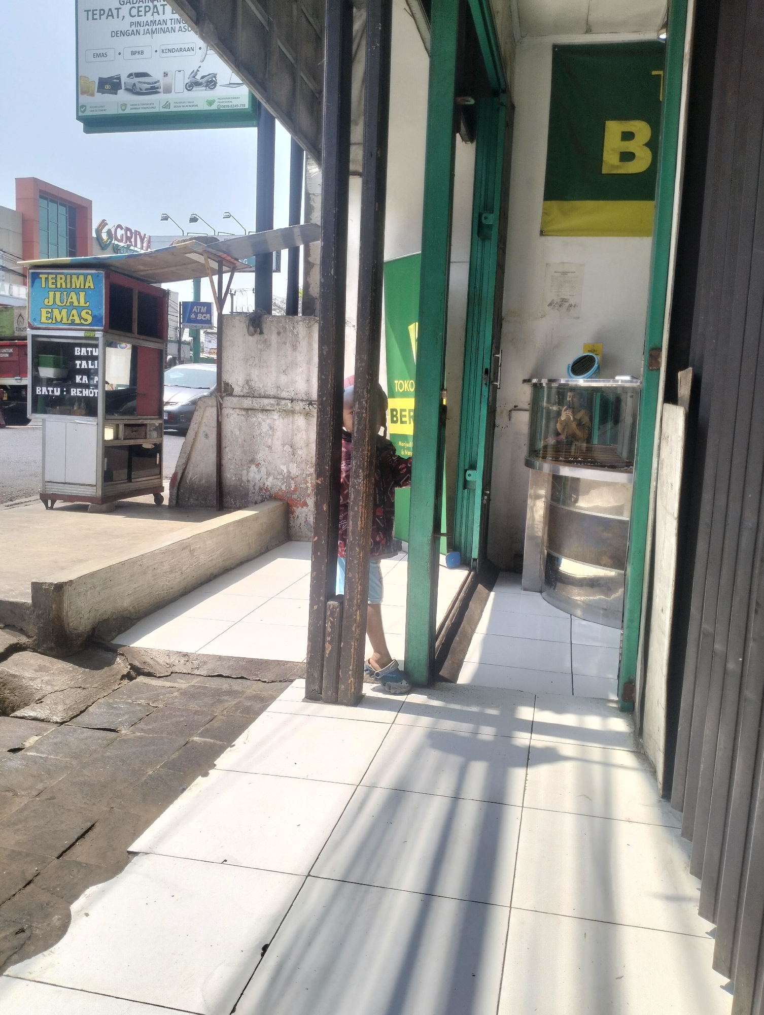 The image shows a street scene in Kel. Pasir Jati, Indonesia, with a focus on a storefront and its adjacent sidewalk. A small, portable cart with the words "TERIMA JUAL EMAS" (Accepts Gold Sales) is visible on the left. Next to it, a sign with information about loans against EMAS, BPKB, and KENDARAAN is displayed. A person, partially obscured, is standing near the entrance of a building, wearing a patterned shirt and shorts. The building's entrance is framed by green metal doors, and inside, a circular display case with a mirror and a blue object is visible. A banner with the letter "B" in yellow on a green and yellow background hangs on the wall inside. The ground is tiled with white squares, and shadows from overhead structures stretch across the pavement. In the background, a building with "GRIYA" written on it and a billboard for "GADAIAN" (Pawnshop) are discernible. A car is parked on the street. The overall environment appears to be a commercial area with signs of daily business activity.