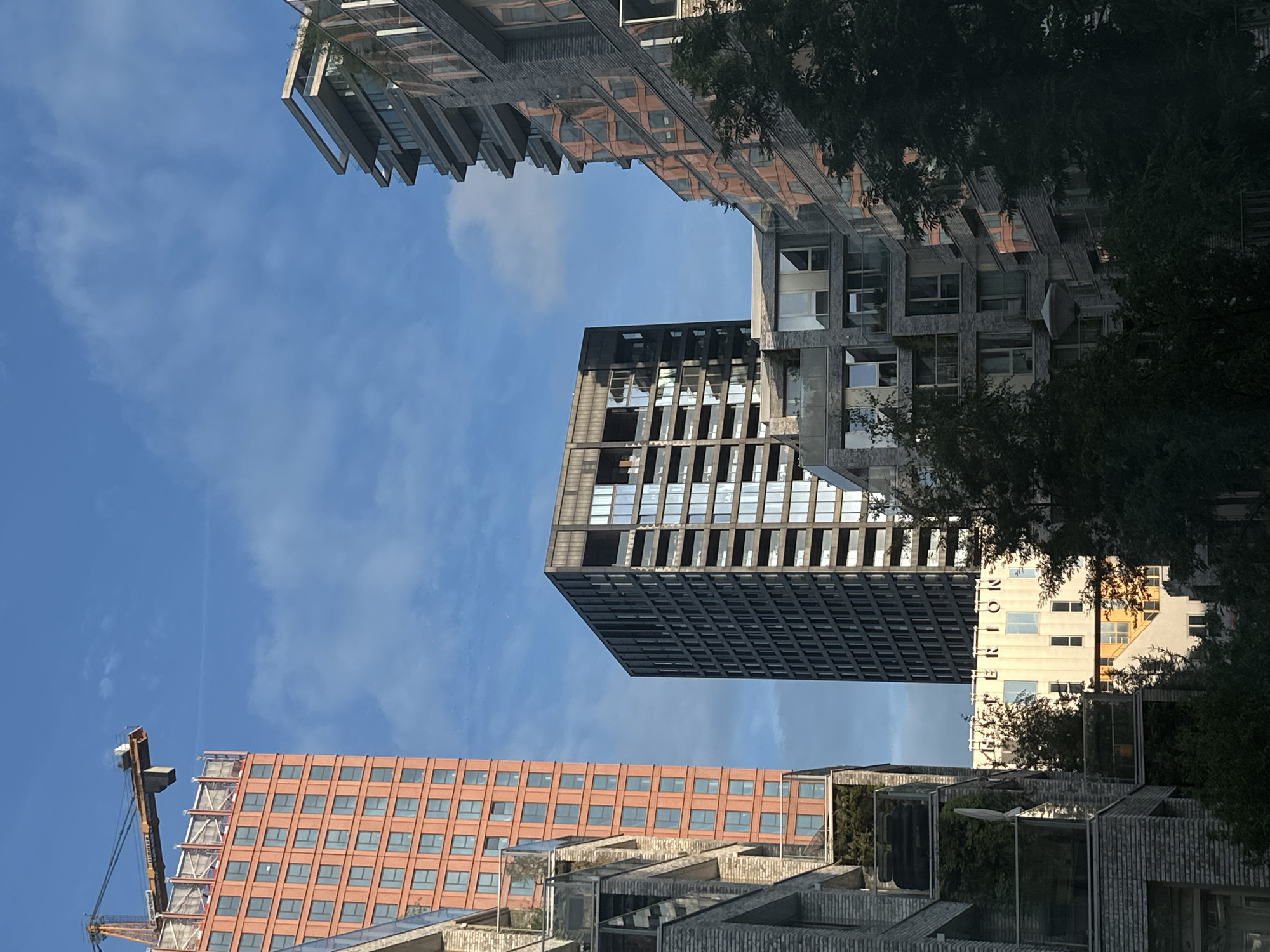 This image depicts a view of residential buildings and a construction crane against a blue sky with some clouds. The buildings are modern in design, featuring various materials such as brick, glass, and metal. The building in the foreground on the left is partially obscured by trees and appears to be made of brick with balconies. To the right, another building with a complex geometric facade is visible, also with balconies. In the center, a darker, rectangular building with many windows stands tall. To the left of this, a tall, orange-bricked building is under construction, topped with a crane. The word "HYPERION" is visible in large letters on a lighter-colored building in the midground. The scene suggests an urban environment, likely a developing area of a city, with a mix of completed and ongoing construction projects. The lighting indicates it is daytime, possibly morning or late afternoon, given the long shadows and the angle of the sun. The image does not contain any people.