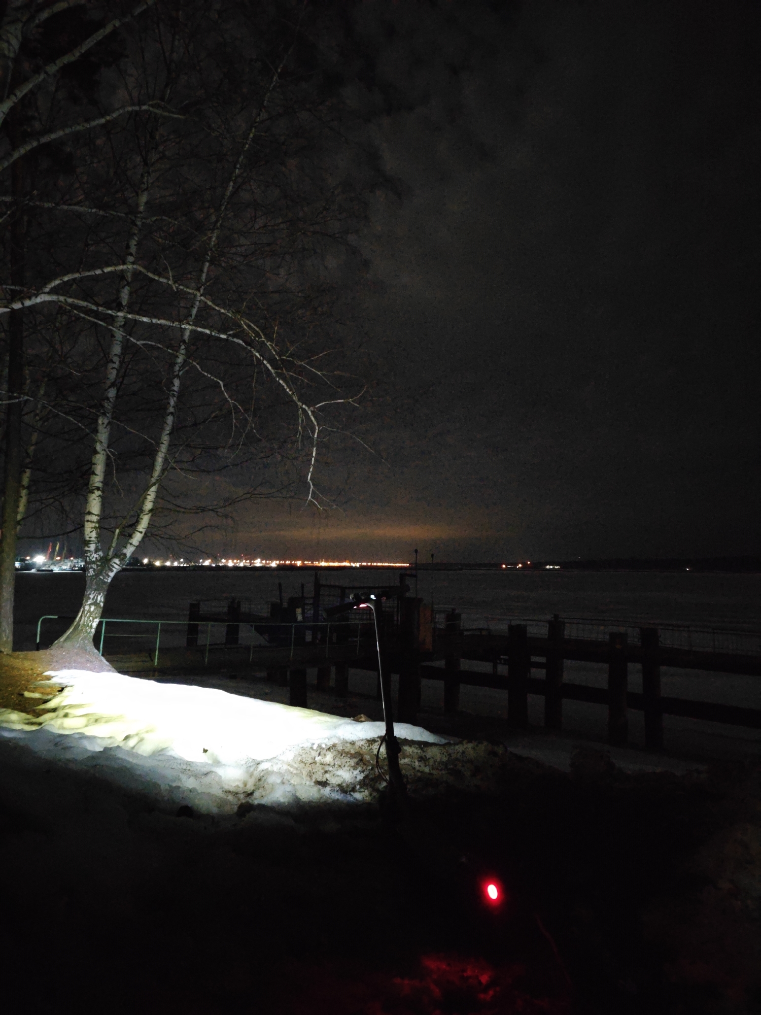 The photograph depicts a night scene in Chaikovsky, Russia, near the Kama River. The foreground features snow illuminated by a bright light source, likely a flashlight or spotlight. A stand of birch trees is visible to the left, their bare branches reaching towards the dark sky. Beyond the snow-covered area, there's a wooden pier extending into the frozen river. In the distance, lights from the city are faintly visible along the horizon. The overall atmosphere is dark and cold. A red light, possibly from a device or signal, is seen in the immediate foreground.