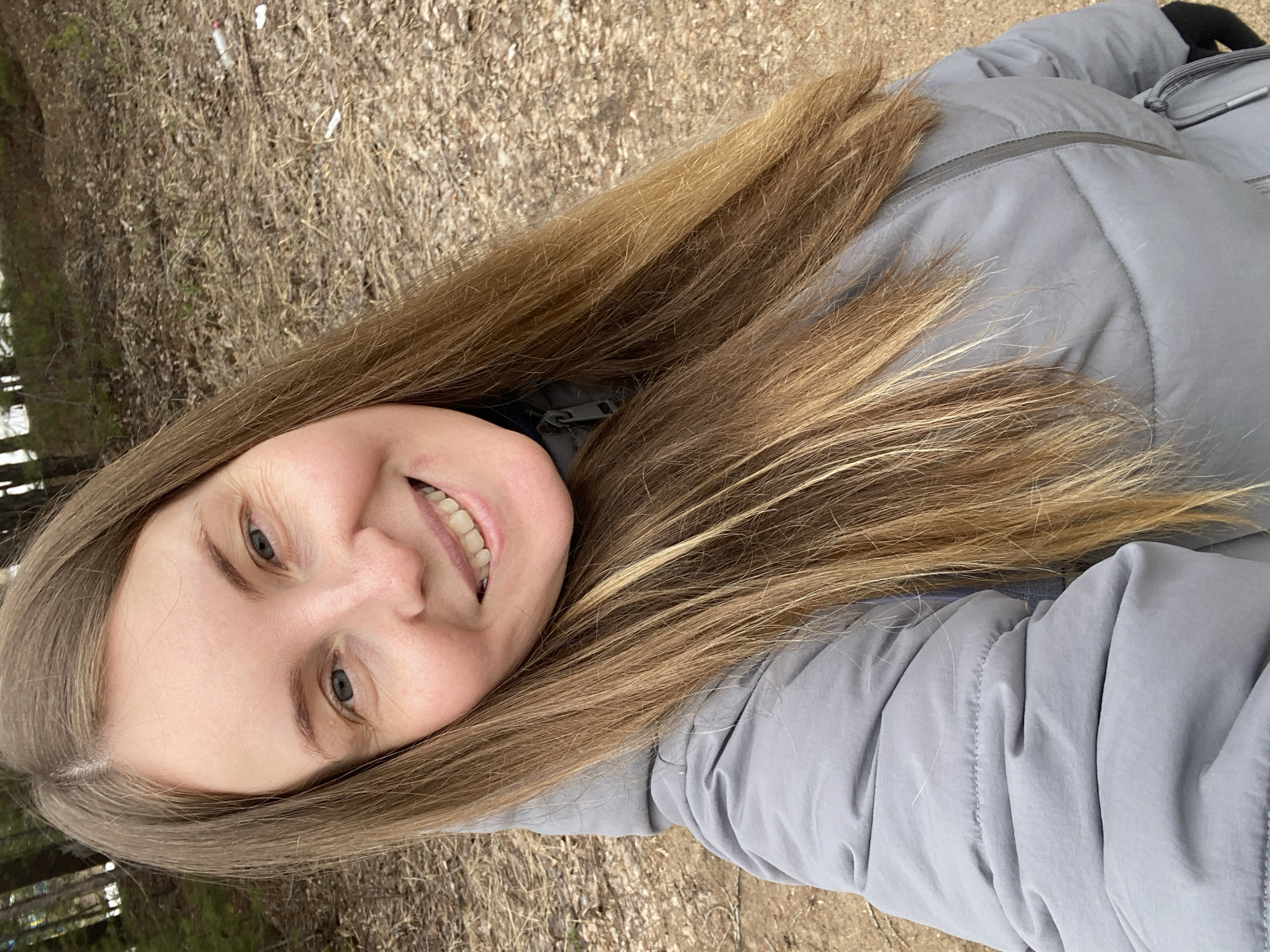 A woman smiles at the camera while wearing a gray jacket. The background appears to be an outdoor location with dry leaves and sparse trees, suggestive of a park or wooded area. The location is not identifiable as Noviy, Russia, from the visual information provided.