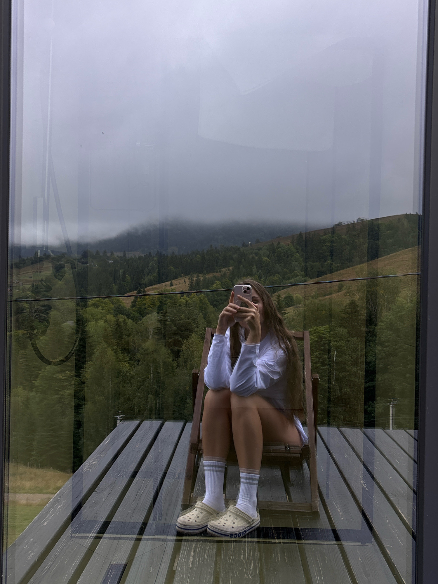 A young woman with long, dark hair is sitting on a wooden chair on a wooden deck. She is wearing a white long-sleeved shirt, white socks with gray stripes, and light-colored clogs with blue accents. She is holding a pink smartphone in her hands and appears to be taking a selfie. The deck overlooks a scenic view of rolling hills covered in lush green trees and mist. In the background, a mountain range is visible, partially obscured by fog. The reflection of the room behind the viewer is visible in the glass. The setting is the village of Krasnyk, Ukraine.