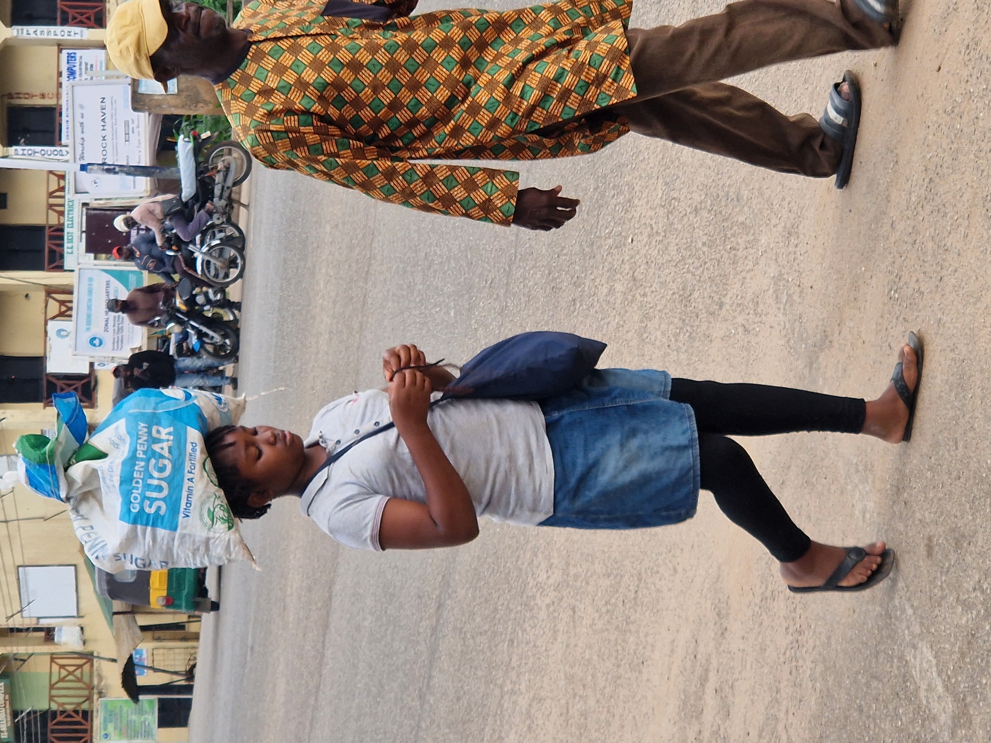 A young person carries a large bag of "Golden Penny Sugar" on their head as they walk down a dusty street in Kabong, Nigeria. The person wears a grey polo shirt, a denim skirt, black leggings, and black flip-flops. To the right, an adult man wearing a yellow cap and a colorful patterned shirt walks in the same direction. Behind them, several motorcycles are parked, and buildings line the street.