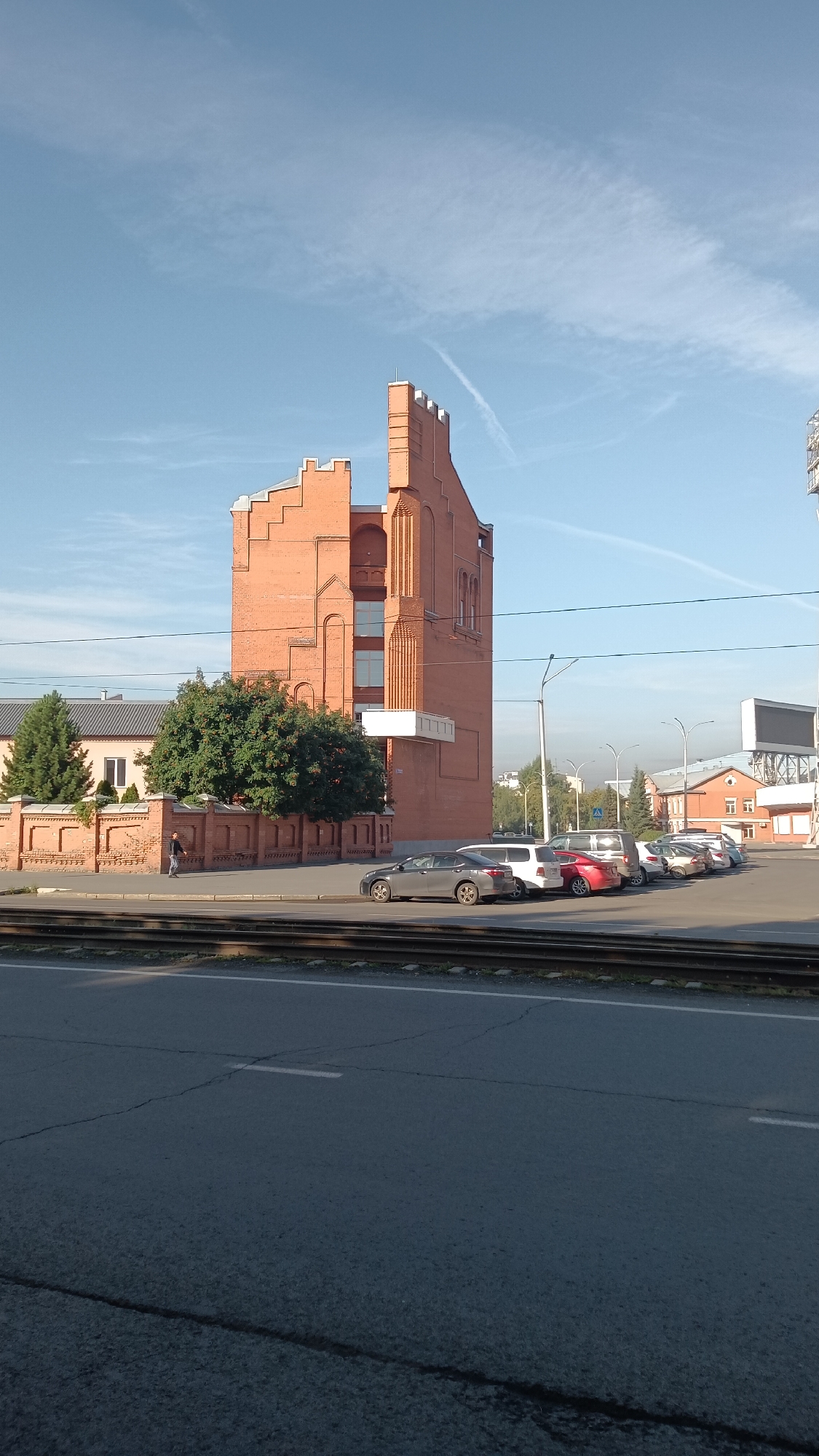 The image shows a street scene in Kemerovo, Russia, on a clear, sunny day. In the foreground, a two-lane asphalt road is visible with some cracking and white lane markings. Train tracks run parallel to the road, separated by a strip of grass and gravel.
To the left of the tracks is a brick wall bordering a property. Behind the wall, a two-story building with a dark grey roof and light pink walls is partially visible. A large, mature green tree stands in front of this building. A single pedestrian, dressed in dark clothing, is walking along the sidewalk next to the brick wall.
Dominating the center of the image is a tall, striking brick building with an unusual, stepped facade and crenellated roofline. The brickwork is a warm, reddish-orange hue. The building features several windows, some of which are arched, and a white rectangular extension on its lower section.
To the right of the brick building, a parking lot is visible, filled with several cars. These include a grey sedan, a white minivan, a red sedan, and several other vehicles parked in a row. Beyond the parking lot, more buildings are visible, including a red-brick structure with white trim and a large, rectangular white sign or screen. Streetlights are also present. The sky is a clear blue with a few wispy white clouds and a faint contrail from an aircraft.