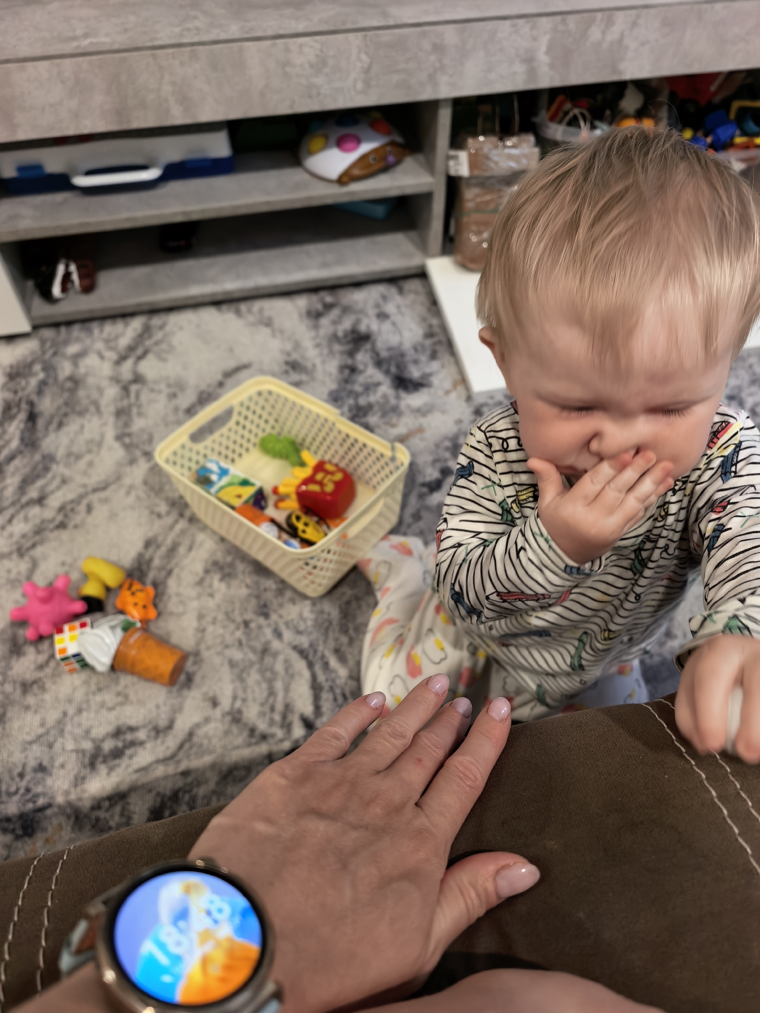 The scene appears to be indoors, likely a home, based on the presence of a carpet with a marble-like pattern and casual objects. A young child, possibly a toddler, stands near a brown couch and is covering their mouth with a hand. They are wearing striped pajamas with various colorful designs. Near the child, on the carpet, there is a small, light-colored basket containing toys and other toys scattered around it. In the foreground, a person’s hand rests on the couch, adorned with a smartwatch displaying an image of a man and a child. A gray cabinet with items stored on its shelves is visible in the background. The location is not evident from the image, nor is the city of Sterlitamak, Russia.