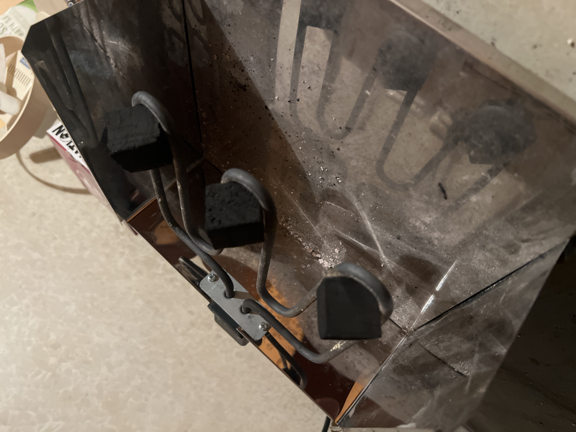 The image shows a used charcoal burner, likely for hookah or incense, with several charcoal briquettes resting on its heating coils. The burner is made of metal and appears to be covered in ash and debris. The background is a textured, light-colored surface, possibly a wall. The scene is indoors, and there is no indication of the specific location in Blagoevgrad, Bulgaria.