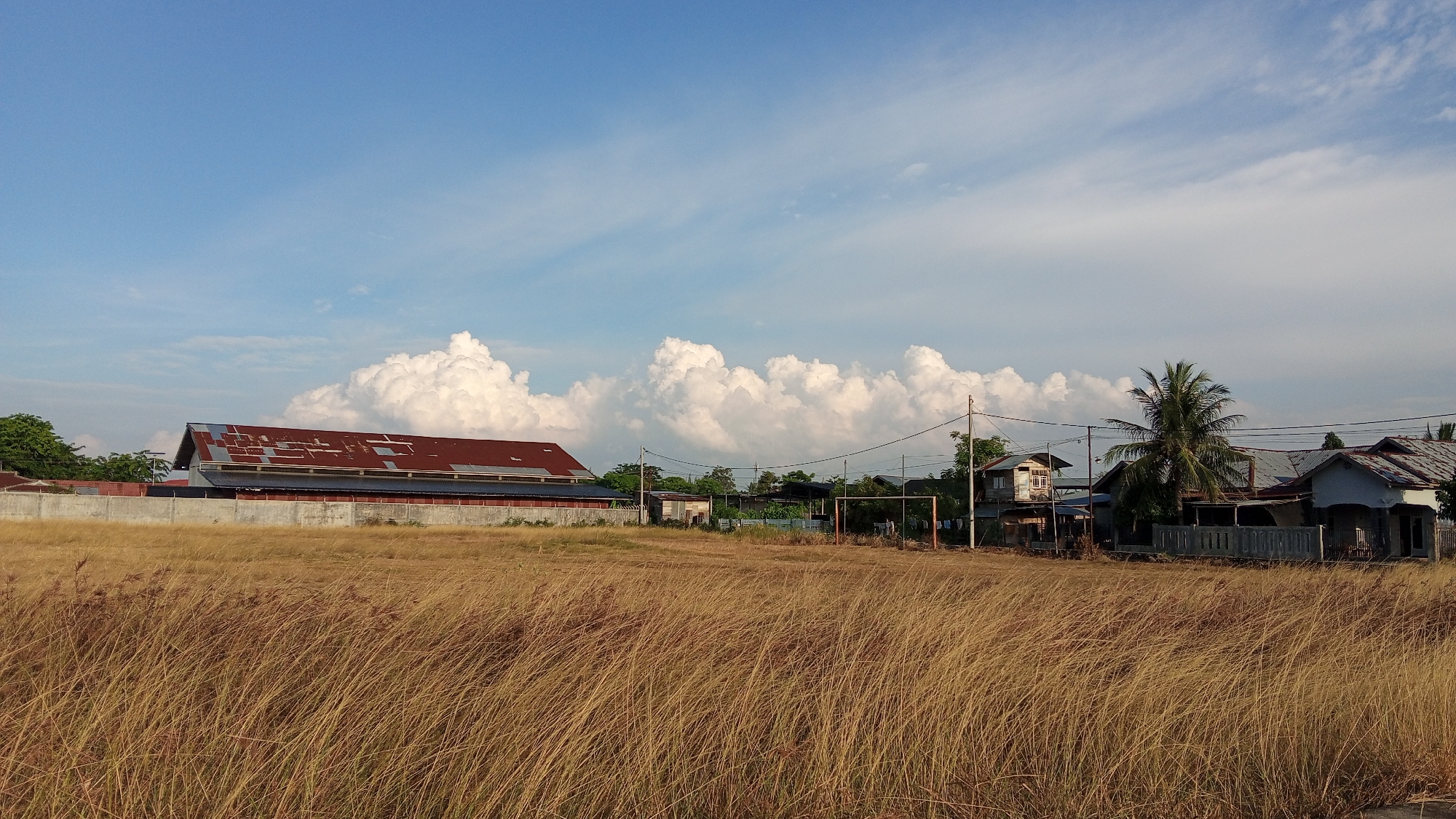 The image depicts a dry, grassy field in front of a cluster of buildings under a bright blue sky with scattered white clouds. On the left, a long, rectangular building with a rusty, corrugated metal roof stands behind a concrete wall. To the right of this building, a small clearing with a soccer goal is visible, along with several modest houses with metal roofs. A tall palm tree stands among the houses. The scene appears to be a rural or semi-urban area in Geulanggang Baro, Indonesia. The overall impression is one of a warm, sunny day.