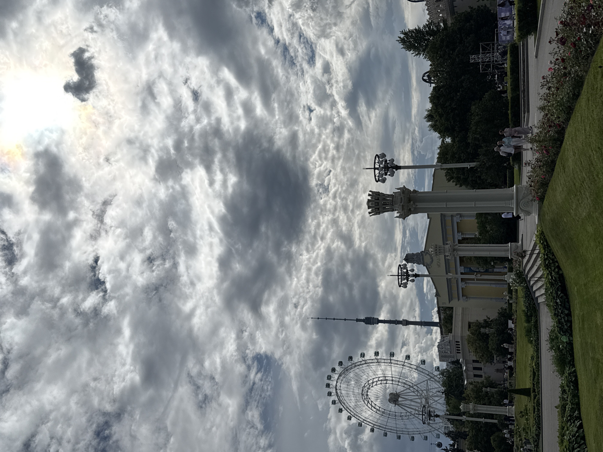 This is a rotated, eye-level, outdoor shot of a park on a cloudy day. The sky is filled with a mix of dark, brooding clouds and bright, white, wispy ones, with the sun partially obscured by a dark cloud. In the foreground to the right, there is a neatly manicured green lawn, followed by a paved walkway lined with flowering rose bushes and manicured hedges. Further back, people are seen walking along the path. Behind the walkway and hedges, there is a large, pale yellow building with ornate white architectural details and columns. To the left of the building, a tall, thin tower pierces the sky, and a large Ferris wheel is visible in the lower right quadrant of the image. Several lampposts with multiple lights are also present, contributing to the park-like atmosphere. The overall scene suggests a daytime visit to a recreational area.