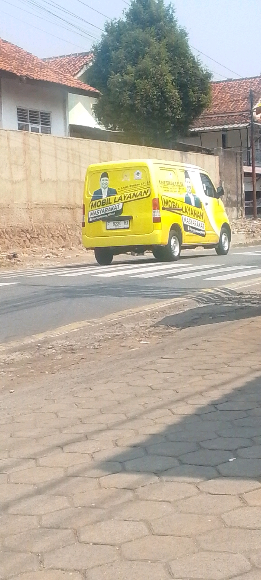 A yellow van with the words "Mobil Layanan Masyarakat" (Community Service Vehicle) and portraits of two men is driving on a road with a pedestrian crossing. The van has an Indonesian license plate that reads "F 8220 HX". The background shows a building with a red tile roof and a large green tree. The road is asphalt and the pedestrian crossing is marked with white stripes. The scene is outdoors in what appears to be a residential area.