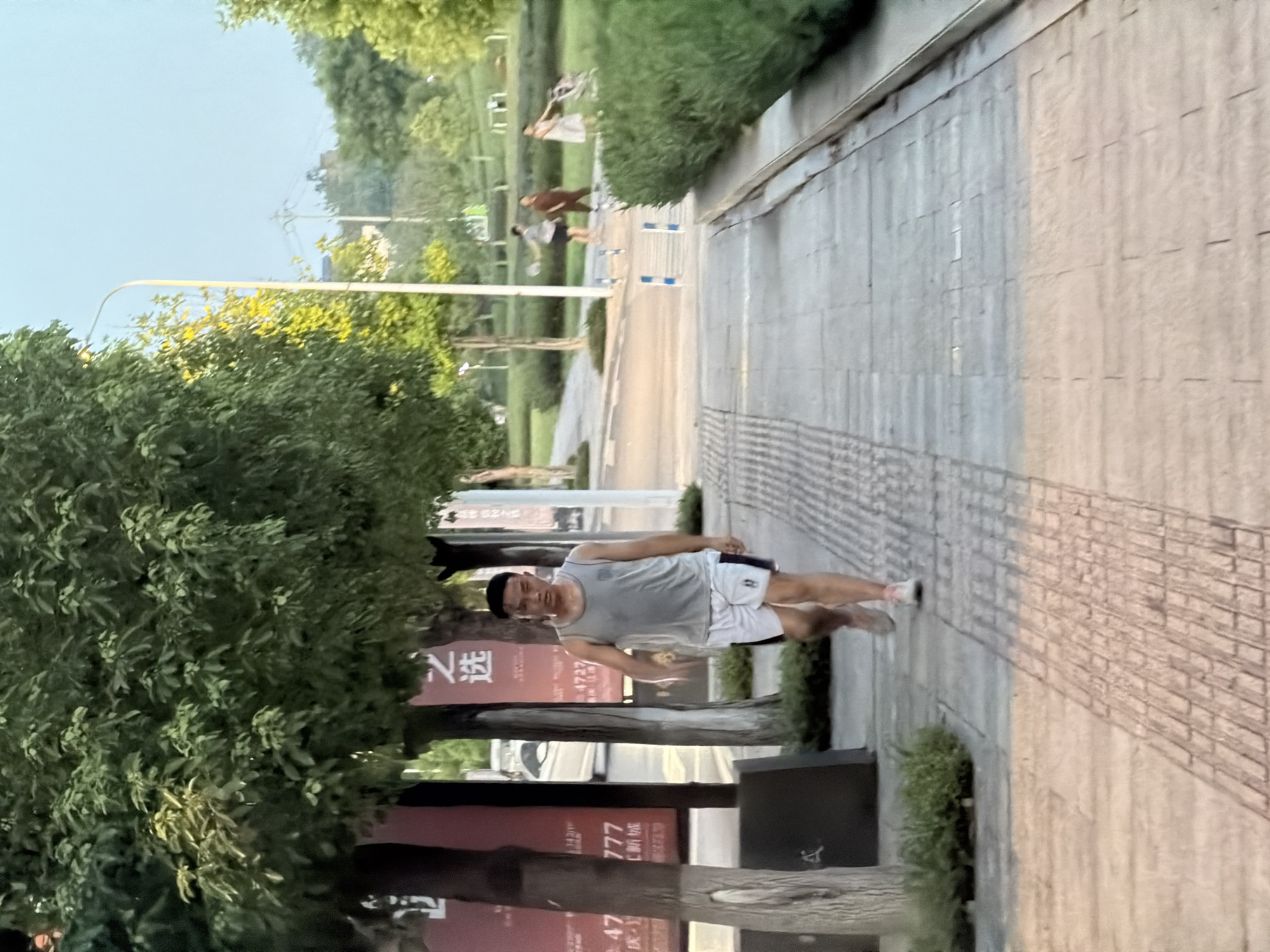 The image shows a young man walking on a paved path in a park-like setting in Jiangjin, China. He is wearing a grey tank top and white shorts with a black stripe. The path is lined with trees and bushes. In the background, other people can be seen walking and pushing a stroller. The sky is overcast. The scene appears to be in the late afternoon or early evening, as indicated by the soft lighting. There are advertisements on the left side of the image.