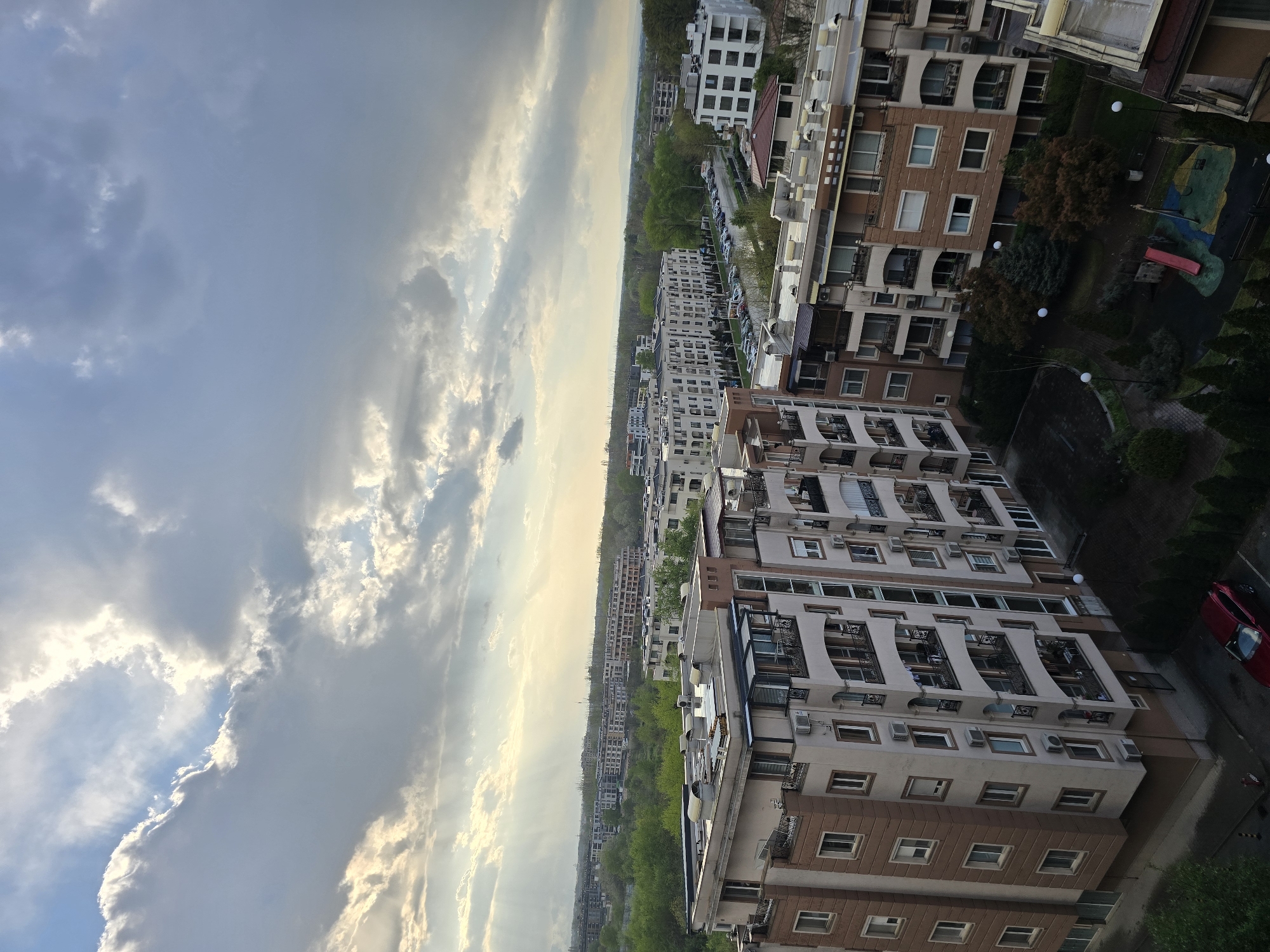 The image shows a cityscape in Unknown, Unknown, taken from a high-angle perspective. Predominantly, residential buildings are visible, with a mix of light-colored facades and darker brown accents. The buildings have multiple stories and feature balconies, windows, and what appear to be air conditioning units. A street runs between the buildings with parked cars. The right side of the picture has a red car parked. A playground is seen on the right side, with green trees. In the distance, there is a hazy view of the city, transitioning into a cloudy sky. The sky is partially covered with clouds.