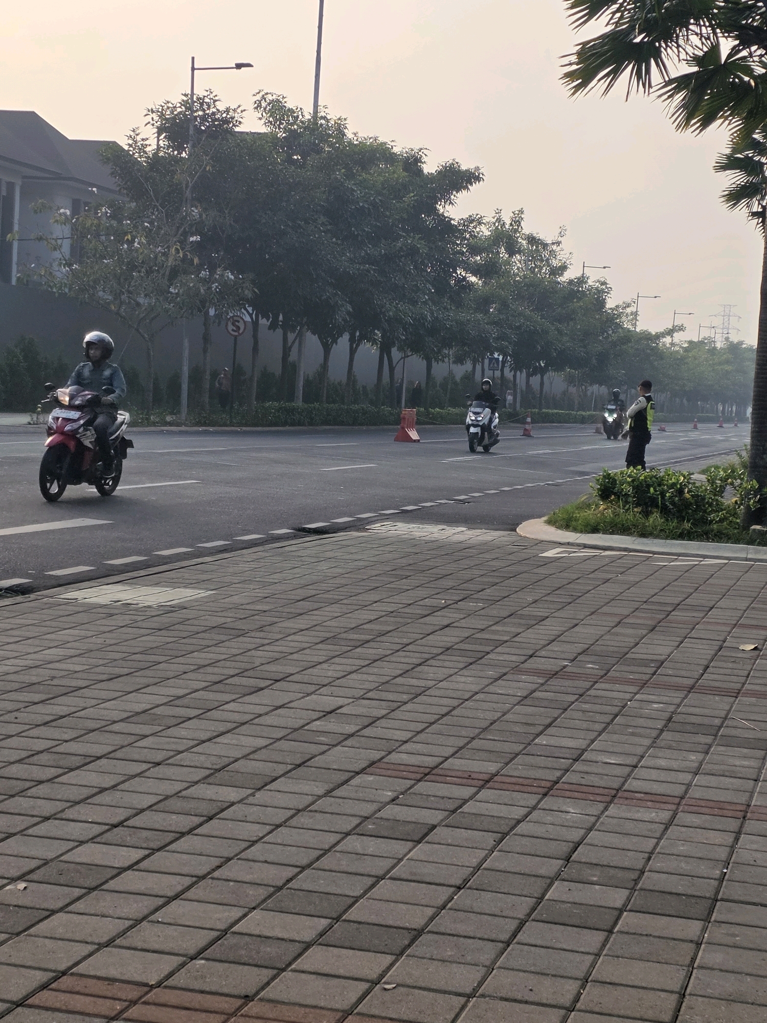 The image depicts a street scene in Bojongsoang, Indonesia, under hazy daylight conditions. In the foreground, a grey paved pedestrian walkway with a repeating pattern of rectangular stones dominates the lower portion of the frame. The street runs horizontally across the mid-ground.
On the street, a person wearing a helmet is riding a red and white motorcycle in the left lane. Further down the road, another person on a white motorcycle is visible in the same lane. In the right lane, a person in a yellow vest and dark pants, possibly a traffic officer or security guard, stands near a lush green bush. Two more motorcycles are seen further in the distance on the right side of the road.
The environment includes a row of trees lining the right side of the street, with more trees and a building visible in the background on the left. Several orange traffic cones are placed along the roadside. Power lines and a distant transmission tower are visible in the hazy sky. The overall atmosphere suggests an early morning or late afternoon due to the soft lighting and slight haze.