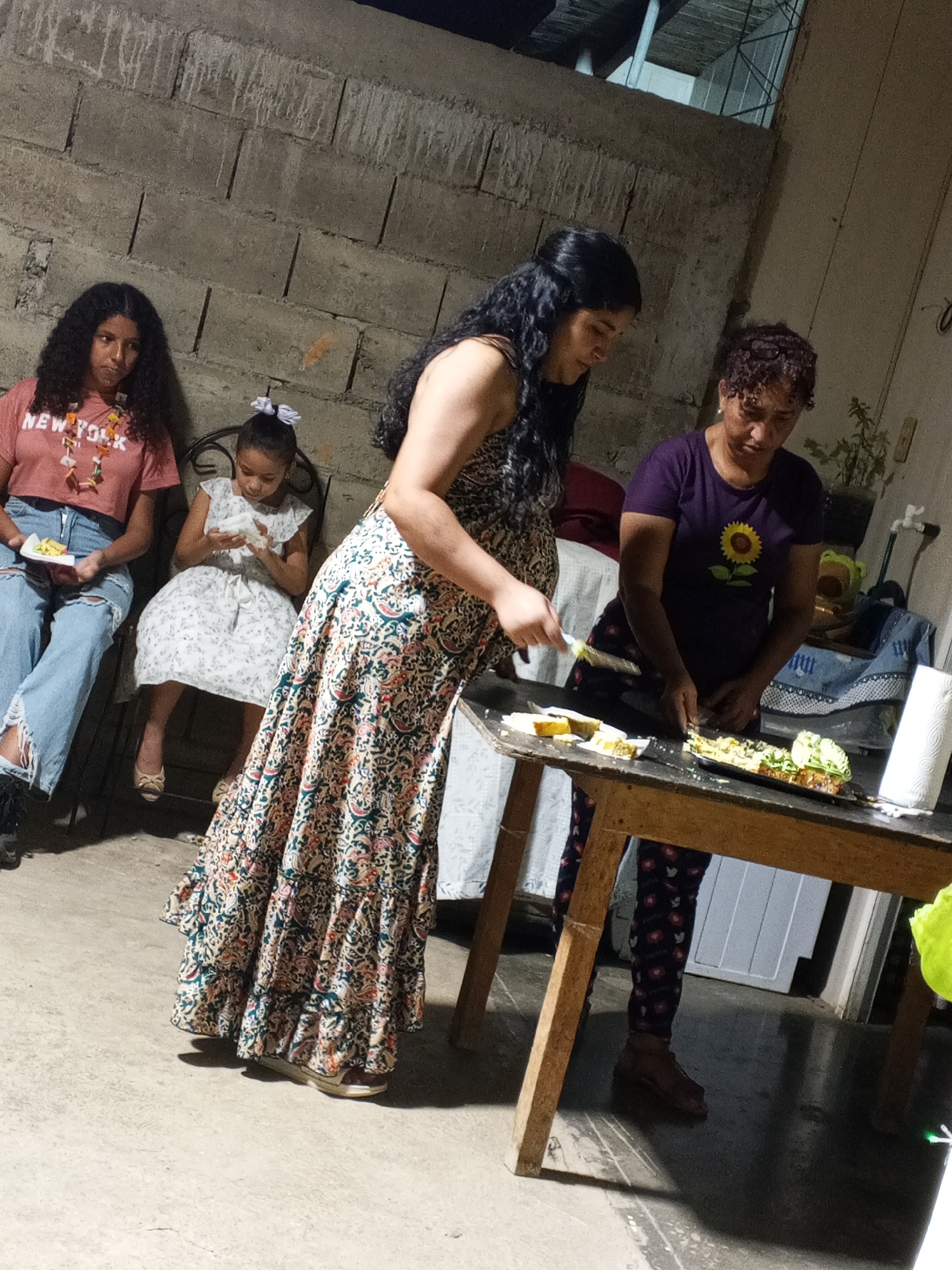 The image shows an indoor scene with four people. On the left, a young woman sits on a chair holding a plate of food, wearing a pink shirt and ripped jeans. Next to her, a young girl in a white floral dress sits on a chair, holding a napkin. Standing and slightly angled to the right is a woman in a long, flowing floral dress. To the right, another woman stands behind a wooden table, cutting food on a plate with a knife. She is wearing a purple shirt with a sunflower print and patterned pants. A roll of paper towels is on the table. The background consists of a cinder block wall on the left and a doorway and window. The location is Unknown, Unknown.