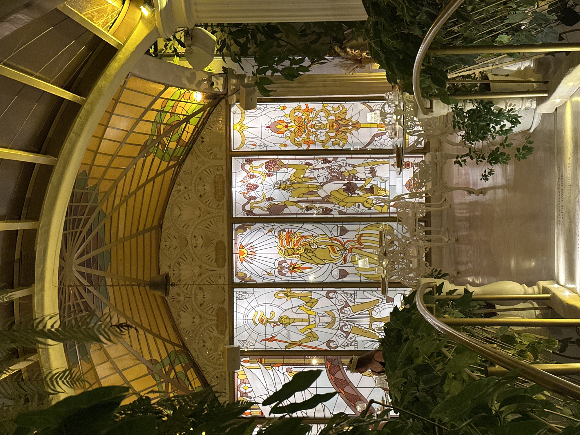 Captured from an overhead perspective, the image showcases an interior space with distinctive architectural features. A prominent stained-glass window displaying motifs reminiscent of Egyptian iconography is a central element. The room also includes a spiral staircase with a gold-colored railing, partly concealed by greenery, and a column topped with a lighting fixture. A window with an arched frame and gold accents further illuminates the space. The scene implies an upscale indoor environment, with details hinting towards a decorative design. This image was taken at The Royal Pitamaha Resort, Ubud District, Indonesia.