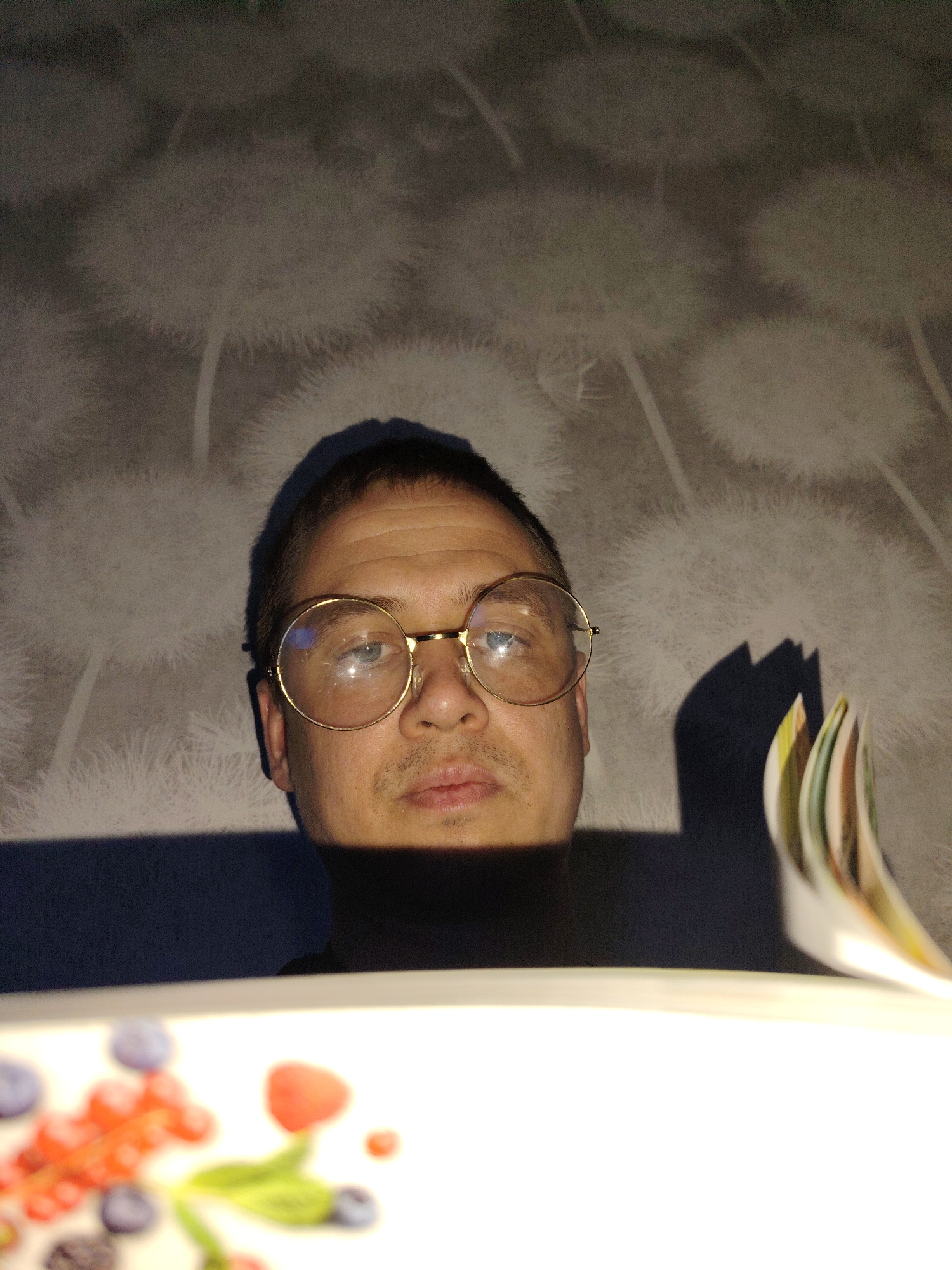 A man wearing gold-rimmed glasses and a black turtleneck is centered in the frame. His face is partially obscured by shadow. He appears to be reading a book or other bound material, as the edge of white pages is visible on the right. A plate with a fruit design is in the foreground. The background is a patterned wallpaper with images that resemble dandelions. While a location in Chaikovsky, Russia has been provided, there is no verifiable information from the image to support this claim.