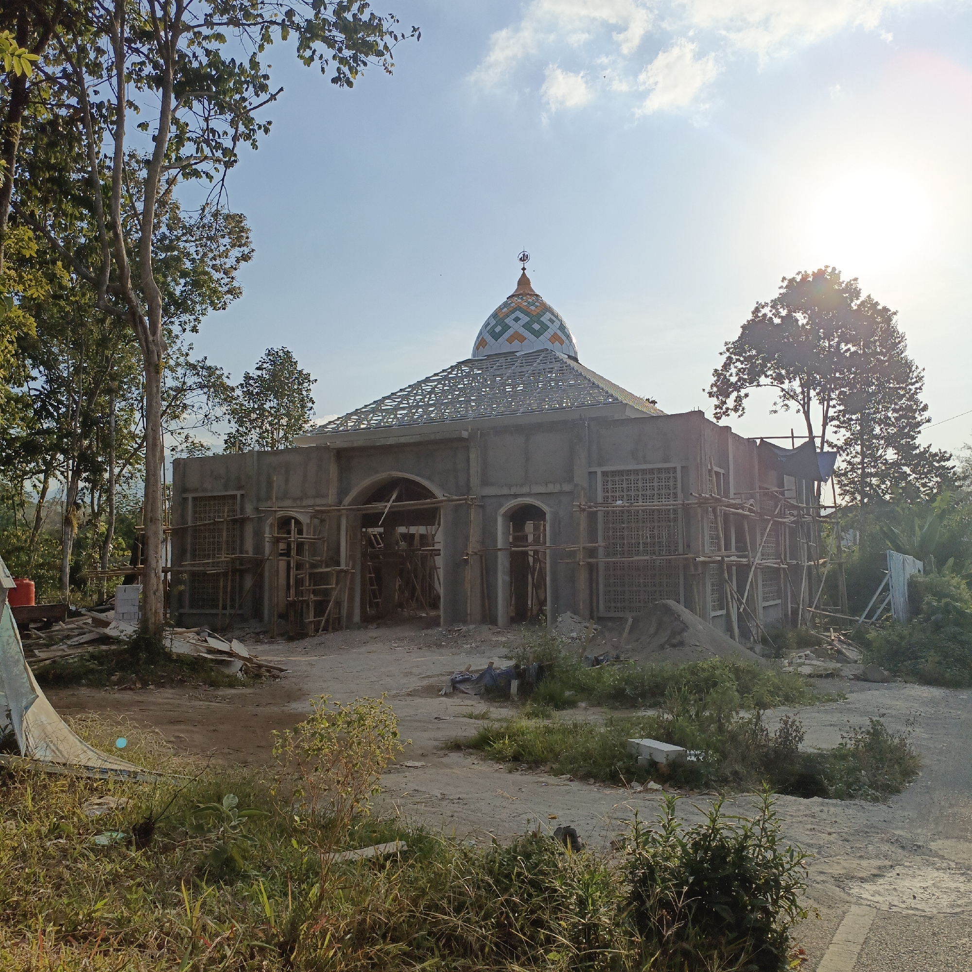 The image depicts the ongoing construction of a mosque in Salatiga, Indonesia, under a clear blue sky with some scattered clouds and the bright sun shining. The mosque, partially completed, features a distinctive dome with a geometric pattern of white, green, and orange tiles, topped with a crescent moon finial. Scaffolding made of wood surrounds the exterior of the building, indicating active construction work. The structure appears to be made of concrete and has arched doorways and windows with decorative lattice work. Piles of sand and construction materials are visible around the site. Several trees, some tall and leafy, are scattered around the mosque, and dense vegetation lines the foreground and edges of the scene. The ground is a mix of dirt and gravel, with some scattered debris, suggesting an undeveloped or rural setting within the city of Salatiga. There are no people visible in the image.