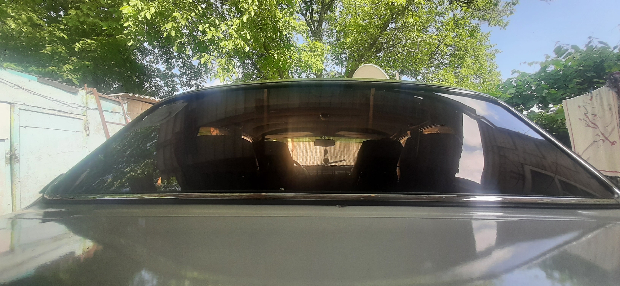 The image shows the rear windshield of a light-colored car, reflecting a yard scene with a wooden fence, trees, and a satellite dish on the roof of a building. Inside the car, the back seats are visible, along with a rear-view mirror hanging from the ceiling. The car is parked outdoors, possibly in a residential area, with sunlight filtering through the leaves of nearby trees. There are no people visible in the image. The location is Yessentukskaya, Russia.