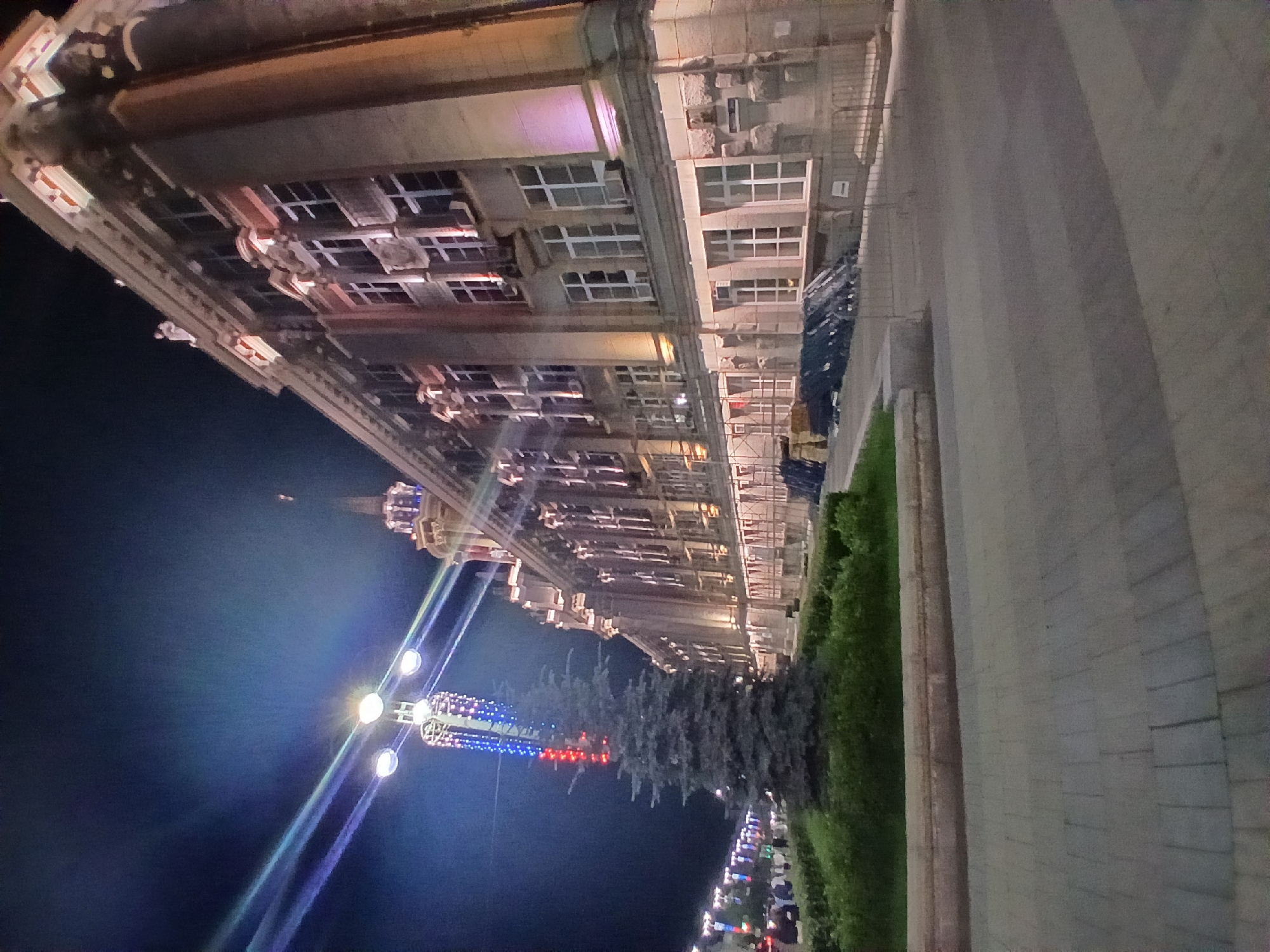 This is a nighttime, slightly low-angle, wide shot of the Yekaterinburg City Duma building, located at 108 Lenin Avenue, Yekaterinburg, Russia. The building is illuminated by external lights, highlighting its classical architectural style with ornate detailing, columns, and multiple floors of windows. A tall, slender fir tree stands in the foreground to the left of the building, situated within a landscaped area with bushes and a stone border. A tall street lamp with multiple bulbs is visible behind the tree, casting light rays into the dark sky. In the background, a street is lined with more lights and people are walking. The foreground shows a paved walkway made of interlocking gray bricks, with a slight curve. Construction barriers and materials are visible near the building on the right side. The overall scene depicts a well-lit public space in front of an important municipal building at night.