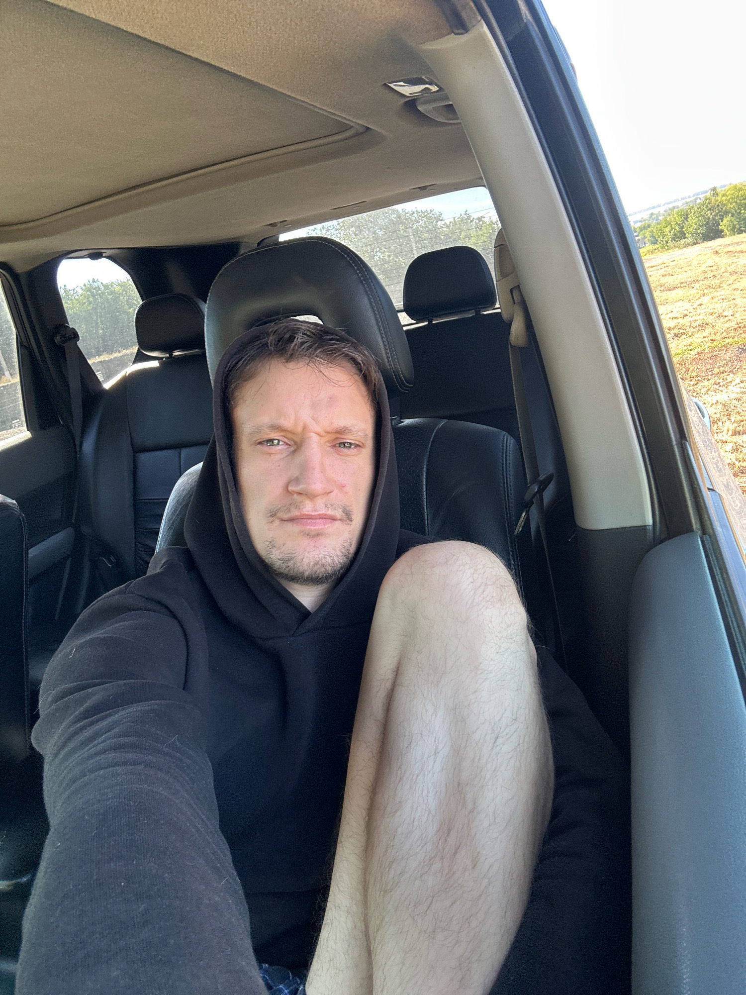 A man is sitting in the driver's seat of a car, looking at the camera. The car's interior is visible, with black leather seats and a sunroof. Outside the car, a field of dry grass and trees can be seen under a bright, sunny sky. The man is wearing a black hoodie and has his legs crossed. His expression is neutral, and he appears to be taking a selfie. The scene is likely outdoors, possibly in a rural area near Makiv, Ukraine, given the natural landscape visible through the car windows.
