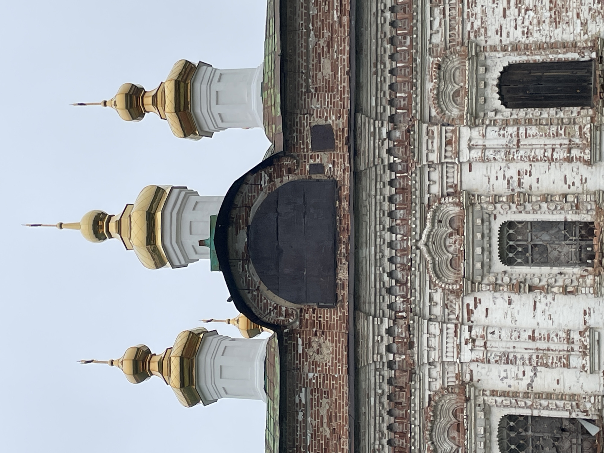The image shows a close-up view of the upper facade and domes of the Church of the Ascension in the village of Kstinino, Tver Oblast, Russia. The church features multiple golden domes with crosses atop white octagonal drums. The brickwork of the building is weathered and in a state of disrepair, with peeling white plaster revealing the red brick beneath. Decorative brickwork and window surrounds are visible. The sky is overcast and grey.