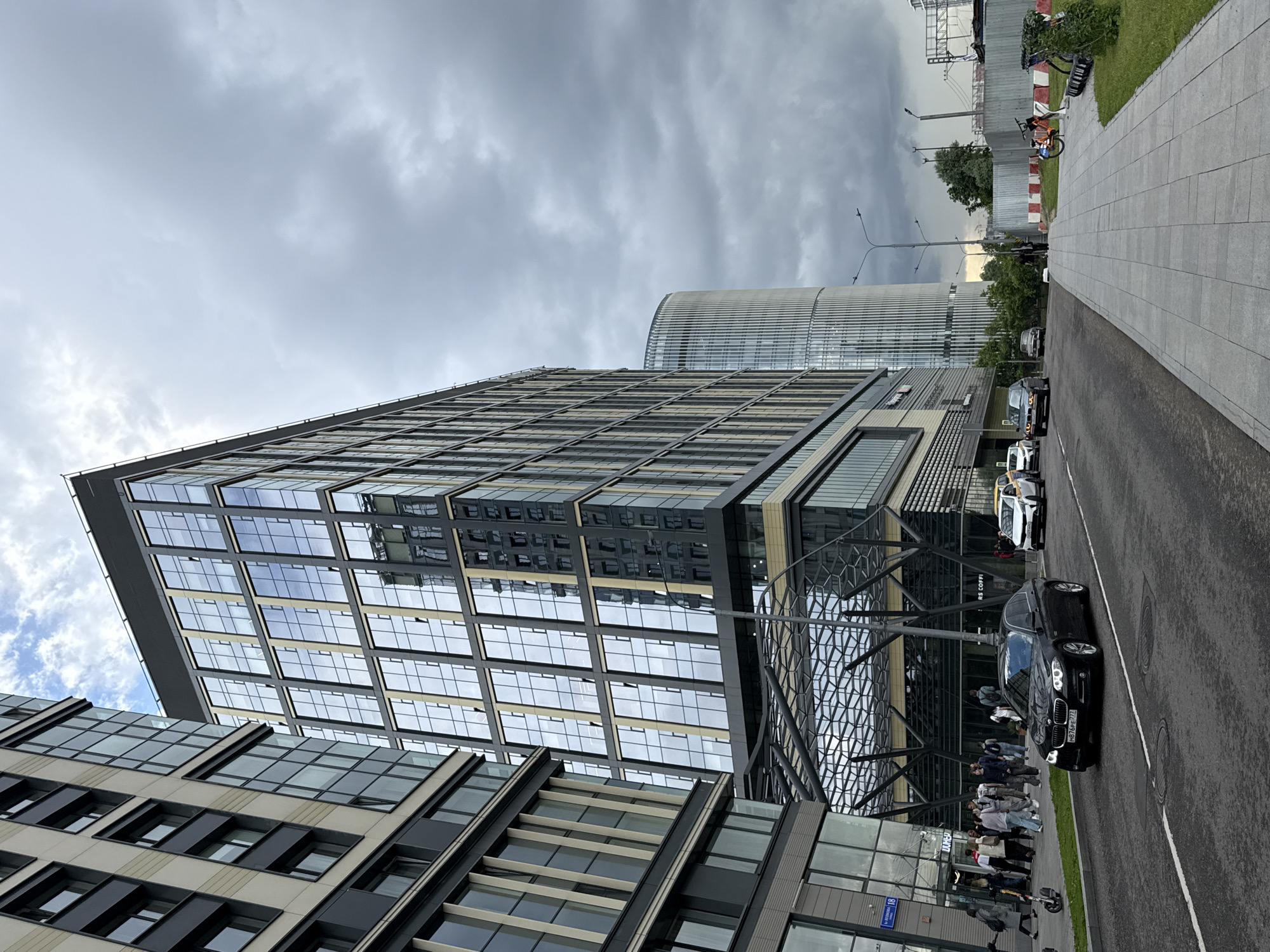 This media file captures a street-level view of a modern urban environment, likely in Moscow, Russia. The scene is dominated by large, contemporary buildings with extensive glass facades that reflect the overcast sky. In the foreground, a black car is parked on the right side of the asphalt road, with a white line marking the edge. A sidewalk made of large, light-colored pavers runs alongside the road.
Several people are visible on the sidewalk, some walking and others standing in groups. Their clothing suggests a casual, everyday setting. A black electric scooter is parked near the sidewalk. Further down the road, more cars are visible, including a white van and several other vehicles. The architecture of the buildings is sleek and geometric, with a prominent building featuring a grid-like pattern of windows. Behind these, a taller, cylindrical building with a ribbed facade rises into the sky.
The weather appears to be cloudy, with a muted, grayish light casting a subdued atmosphere over the scene. The time of day is likely daytime, given the overall illumination. In the background, elements of urban infrastructure, such as power lines and traffic signals, are present. There is also some greenery, including trees and grass, indicating that the urban setting is interspersed with natural elements.
A sign with the text "RS COFFI" is partially visible on one of the buildings. Another sign with Cyrillic text is visible on a building to the left, but the text is too small to be legible. The overall impression is one of a bustling, modern city with ongoing daily activities.