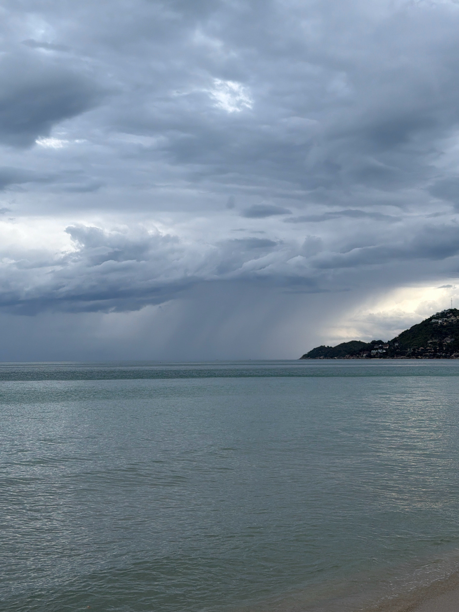 The image depicts a scenic view of the ocean and sky in the city of Ko Samui District, Thailand. The sky is predominantly overcast with dark, heavy clouds, hinting at impending rain. A distinct rain shower is visible in the distance, creating a curtain of precipitation over the sea. The sea itself appears calm, with a muted greenish-blue hue, reflecting the somber sky. To the right, a lush green hillside dotted with houses and structures rises from the water, partially obscured by the dramatic cloud formations. The overall impression is one of a moody, atmospheric coastal landscape. There are no people or animals visible in the frame.