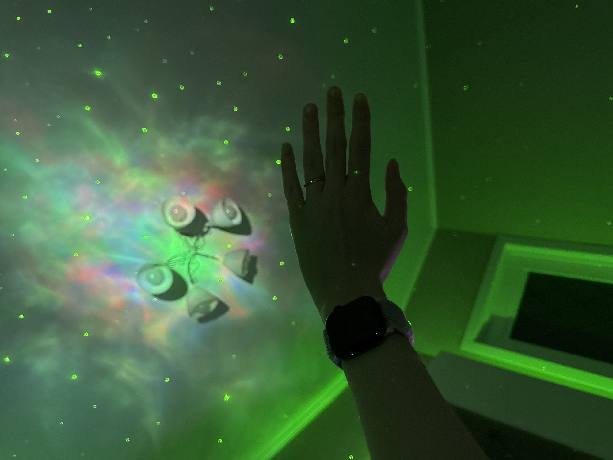 The image displays an indoor scene illuminated by an elaborate light projection on what appears to be a ceiling or wall. Numerous small, distinct green light dots, resembling stars, are scattered across the surface. These green points are integrated with swirling, multi-colored light patterns, primarily green, but also incorporating hues of red, blue, and purple, creating an effect reminiscent of a nebula or an aurora borealis.
In the foreground, a person's right arm and hand are extended upwards into the frame. The hand is depicted with a ring on the ring finger. A smartwatch with a dark screen and a light-colored, possibly woven, strap is worn on the wrist. The arm and hand are largely shadowed, providing a contrast against the bright projections.
On the left side of the image, a light fixture, possibly a chandelier, with several bell-shaped shades is visible, partially obscured by the projected lights. In the bottom right corner, a rectangular architectural element, such as a window or door frame, is prominent with a distinct glowing green outline. The setting suggests a darkened room, emphasizing the vibrant light display. The activity appears to be the person reaching towards or interacting with the projected lights, possibly in a moment of observation or wonder. No text is visible in the image.