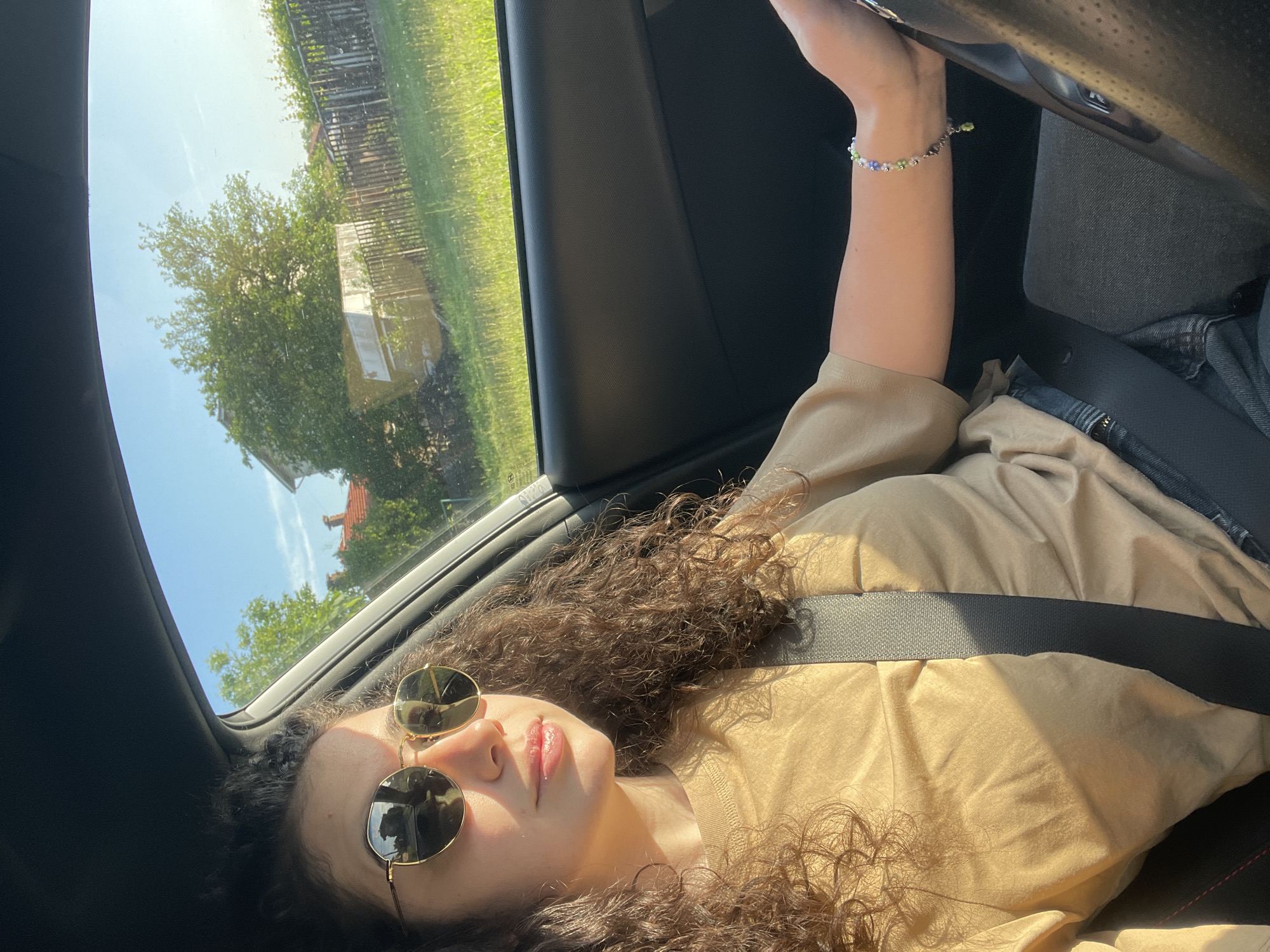 A woman with curly dark hair is resting in the passenger seat of a car. She is wearing sunglasses and a beige shirt, secured with a seatbelt. Her left arm is raised, holding onto a part of the car's interior, and she's wearing a beaded bracelet. Outside the window, trees and a building can be seen, suggesting a suburban or rural area. The location cannot be determined to be Sofia, Bulgaria from the provided information.