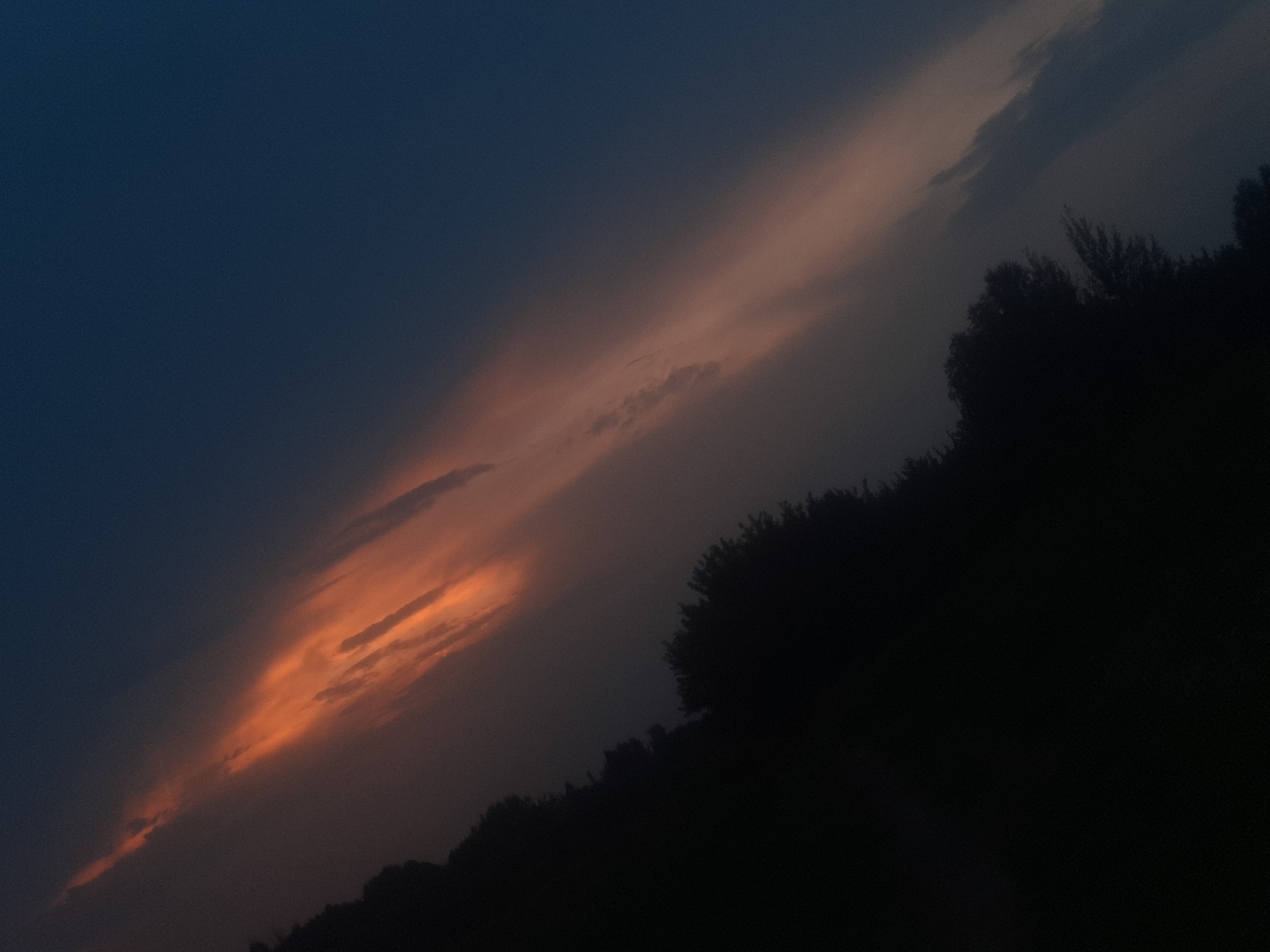 The image shows a diagonal band of orange and pink clouds against a dark blue sky. The lower left portion of the image is dominated by a silhouetted, dark mass of trees, suggesting a rural or forested area. The lighting implies either sunset or sunrise. No people or specific city landmarks are visible. The image appears to be a photograph of the sky and tree line.