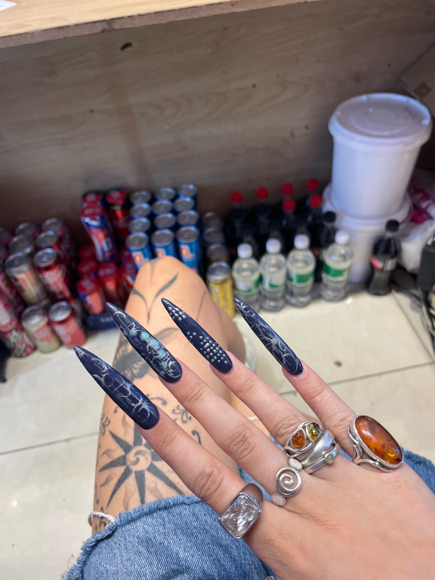 The image shows a close-up of a person's hand and arm, adorned with elaborate navy blue stiletto nails featuring silver intricate designs. The person is wearing multiple rings: a large, oval amber ring on the ring finger, a coiled silver ring with small white stones on the middle finger, a ring with two colored stones (orange and yellow) and a small green stone on the index finger, and a large, square-cut clear gemstone ring on the pinky finger. The person's arm, partially visible, has a large tattoo of a compass rose. They are wearing ripped denim jeans.
In the background, rows of canned drinks (some appearing to be Coca-Cola and possibly other brands) are stacked on shelves made of light-colored wood. Behind the cans, several plastic bottles of water and dark liquid, likely sodas, are lined up. A large white plastic container with a lid is also visible. The environment suggests a storage area or a stock room, possibly within a small shop or convenience store.
There is no information in the image that can definitively place the location in Raanana, Israel. The scene is forensic in nature, presenting objects and a subject without additional context.