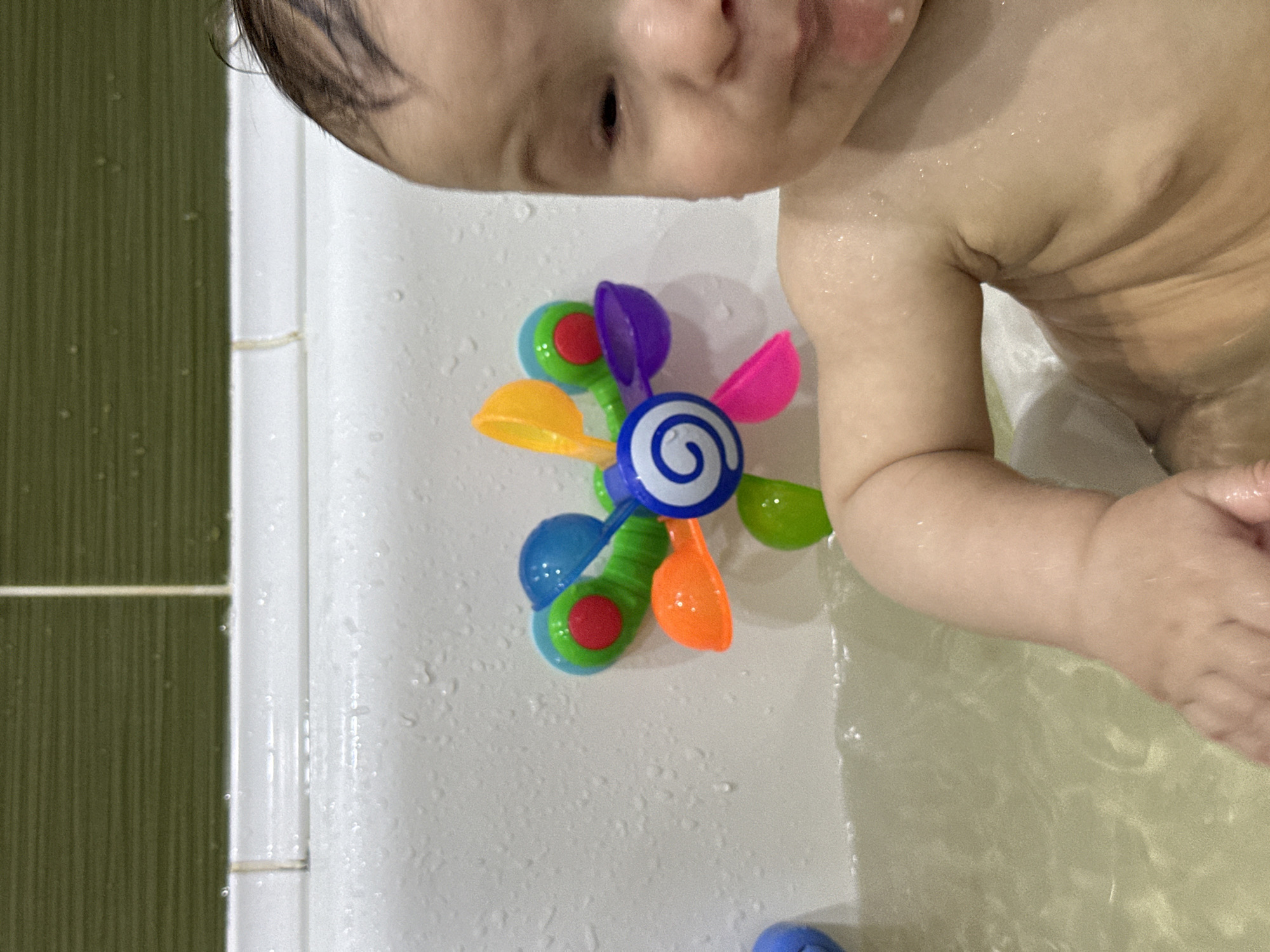 The image shows a baby in a bathtub playing with a colorful suction cup toy. The toy is attached to the white bathtub wall. The baby's head and shoulders are visible. The environment is a bathroom, with green tiles visible on the left.