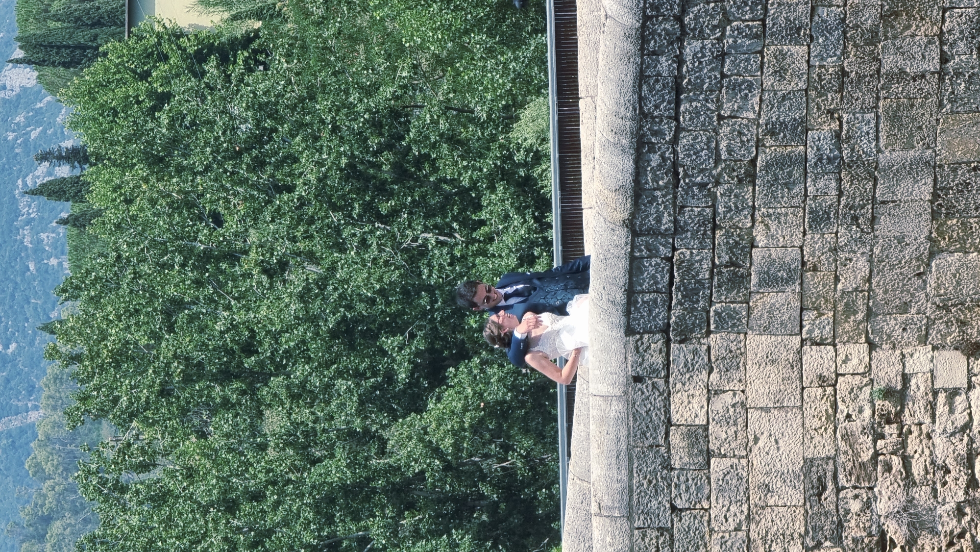 A couple, a bride in a white dress and a groom in a dark suit, stand on a stone bridge in Besalú, Spain. The groom has his arm around the bride. They are looking at each other and smiling. The bridge is made of large, rough-hewn stones. Behind them is a dense forest of green trees, and in the distance, a mountain range. The sky is clear and blue.