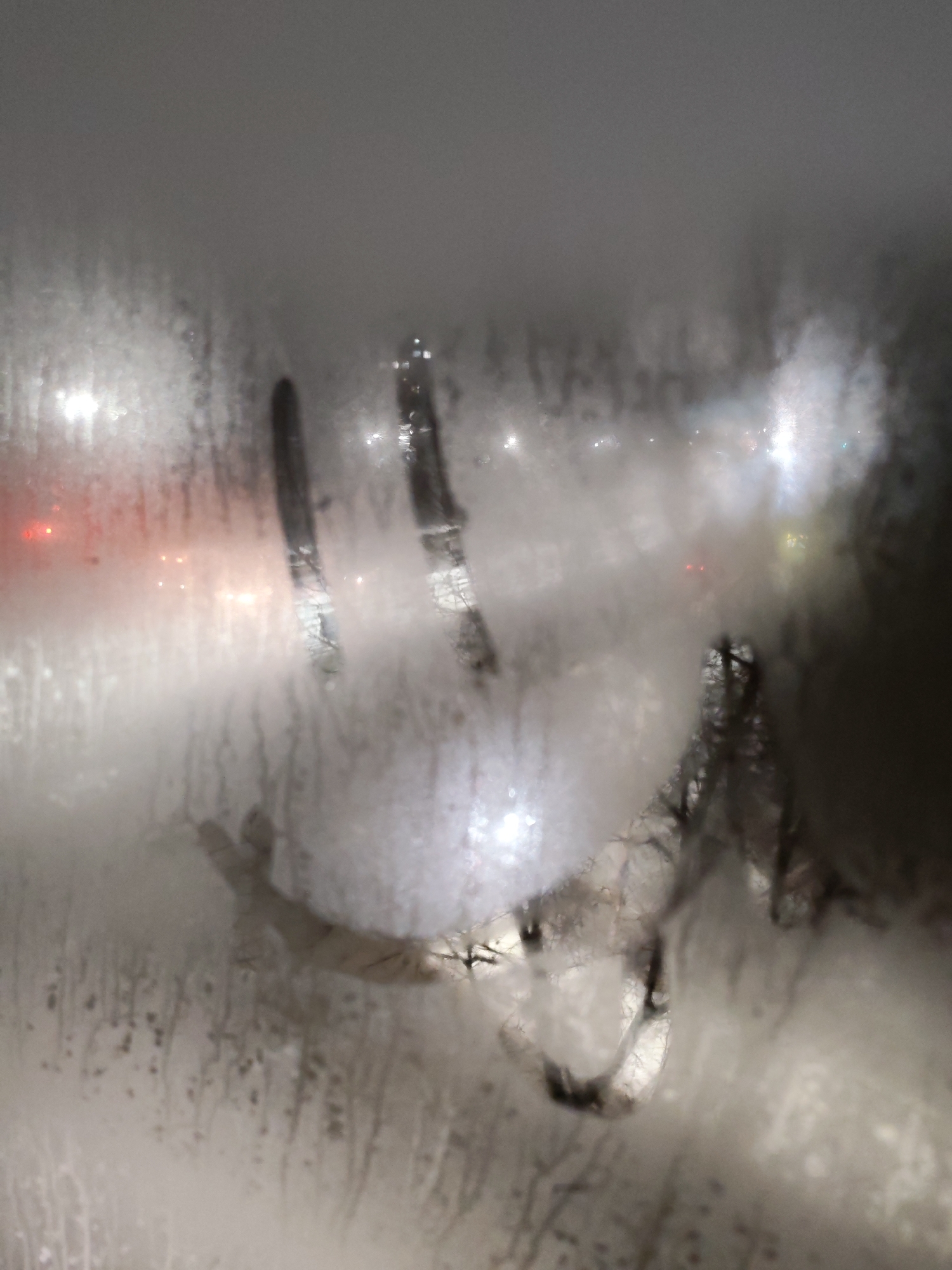 The image shows a foggy window with a smiley face drawn on it. Outside the window, blurred lights suggest a nighttime urban scene. The lights are likely from streetlights and possibly car headlights. The scene is in Izhevsk, Russia, but specific details of the location within the city are not discernible from the image. The smiley face appears to be drawn by a finger. The foreground includes streaks and droplets of condensation running down the glass.