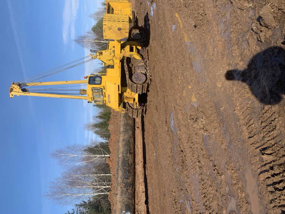An outdoor industrial scene in Sosnovka, Russia, depicts a construction site under a clear blue sky. A large, yellow, tracked pipelayer, identified by the "ЧЕТРА" (CHETRA) manufacturer's label, is prominently positioned slightly to the right of the frame's center. This heavy machinery features a tall boom extending upwards and forwards, equipped with visible cables and pulleys, and a counterweight assembly on its rear. The pipelayer's tracks are heavily coated with reddish-brown mud. To the left of the machinery, a long section of a large-diameter, black-coated pipe, bearing a white 'S' or '5' marking, lies horizontally on the ground, partially settled into what appears to be a shallow trench or prepared bed. The foreground and middle ground consist of disturbed, reddish-brown earth, heavily churned into deep mud with numerous track indentations and standing water puddles. In the background, a line of bare deciduous trees with white bark stands behind a mound of excavated earth, backed by a dense coniferous forest. The shadow of a person, likely the photographer, is cast diagonally across the lower right foreground. The scene indicates ongoing pipeline construction activities under sunny conditions, with the bare trees and muddy ground suggesting late autumn, winter, or early spring.