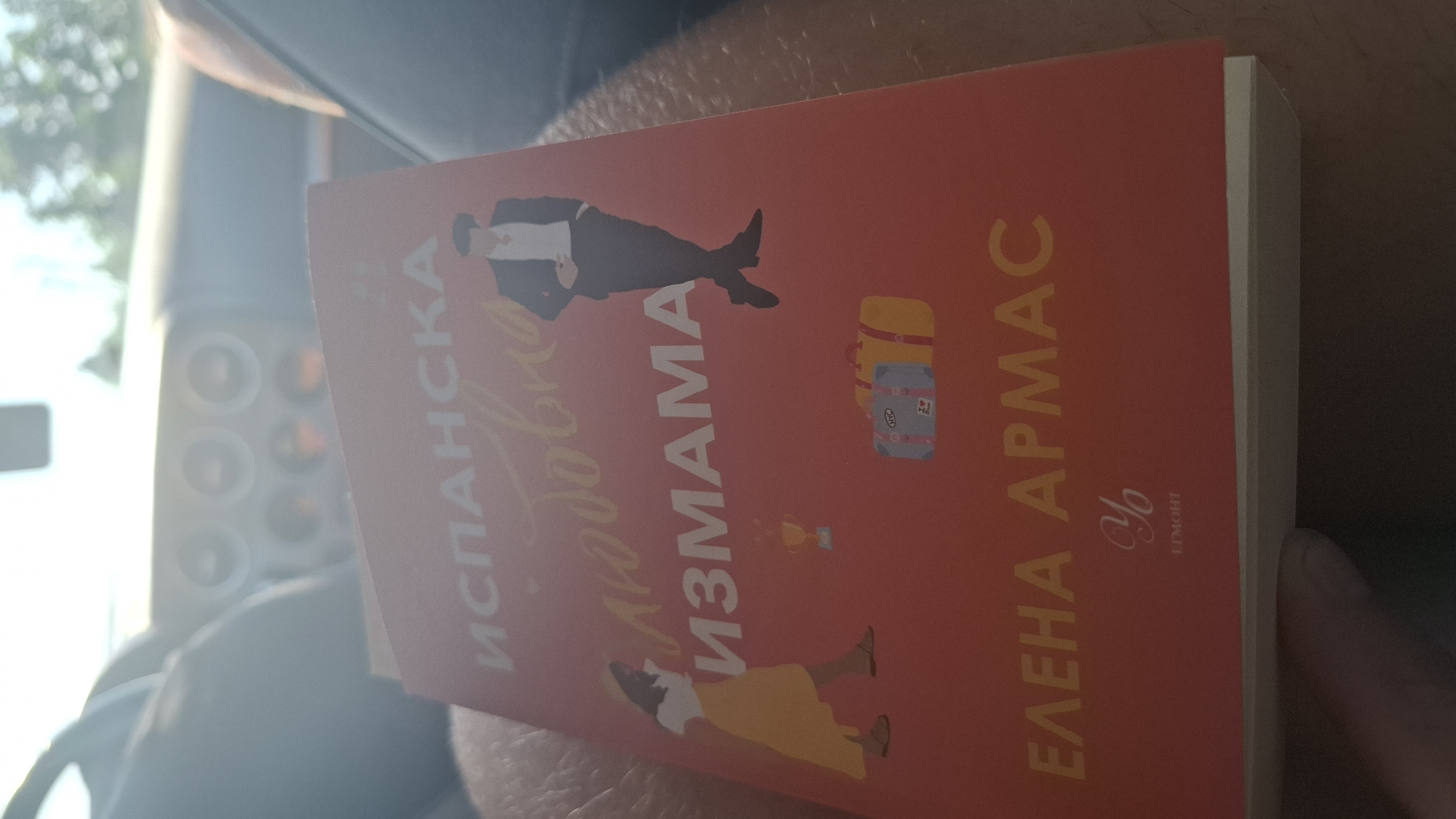 The image shows a close-up of a book with a red cover. The book is titled "ИСПАНСКА ЛЮБОВНА ИЗМАМА" which translates to "Spanish Love Deception". Below the title, there are illustrations of a woman in a yellow dress and a man in a suit, along with two suitcases and a trophy. The author's name, "ЕЛЕНА АРМАС", is printed in yellow at the bottom of the cover. A stylized logo appears below the author's name. The book is held by a person's hand, and the background suggests the interior of a vehicle, possibly a car, with a blurred dashboard and window. The lighting indicates it is daytime. The context of Goren Chiflik, Bulgaria, is not evident from the visual information in the image itself.